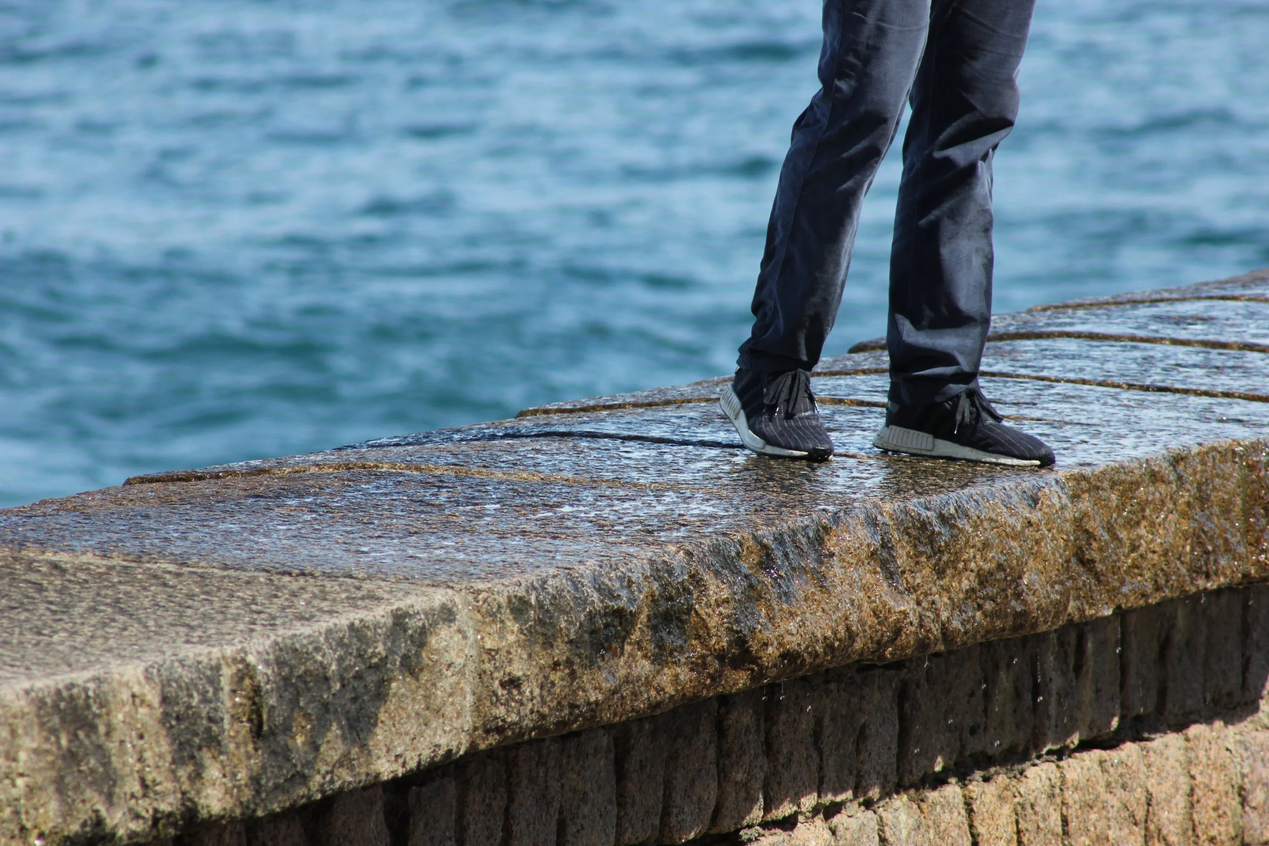 Close-up of a person's legs and feet wearing black sneakers and black pants, standing on a stone pier by the water.