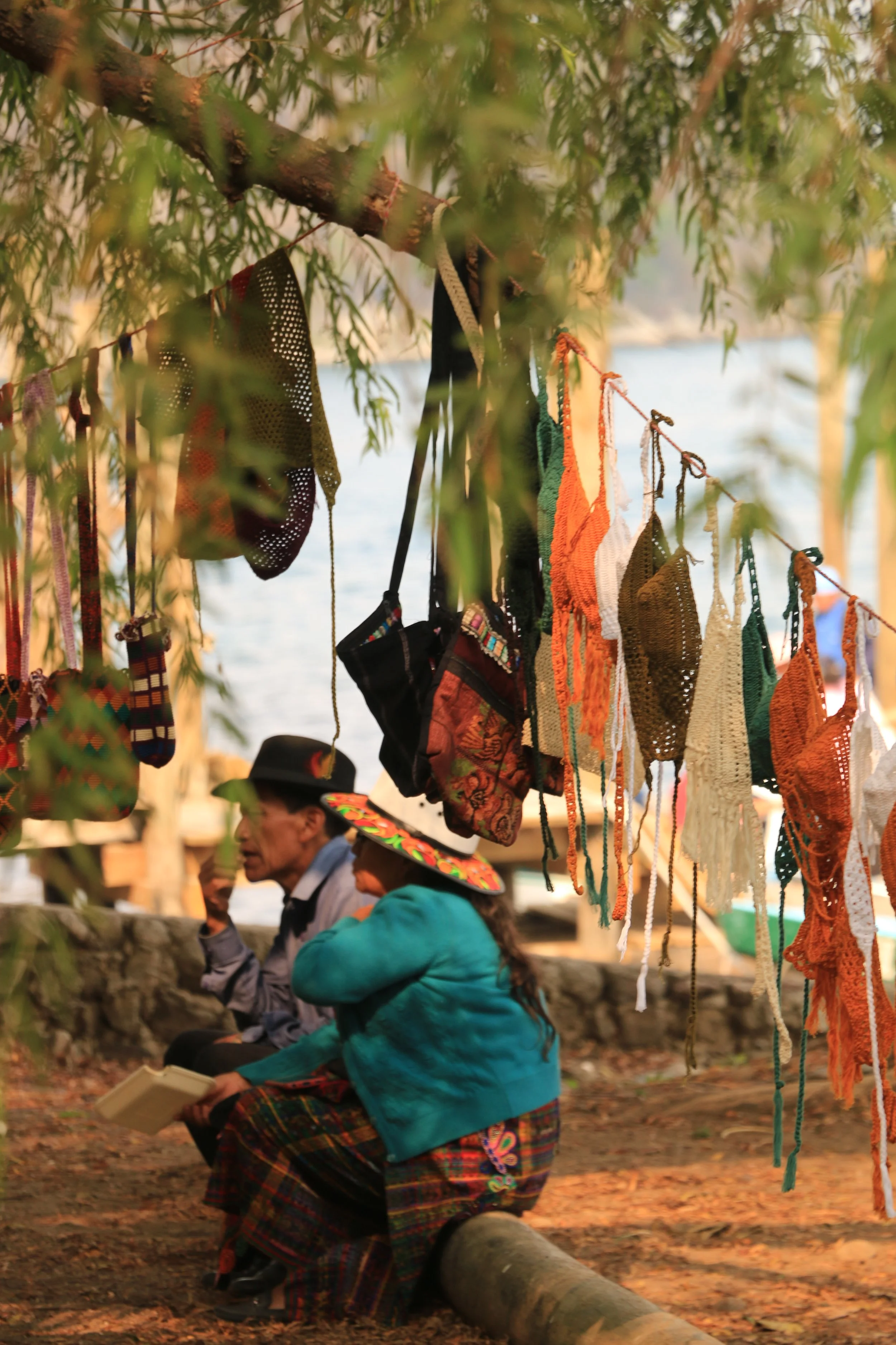 Two people sitting outdoors near a body of water, with colorful knitted and crocheted items hanging from a line above them, and trees in the foreground.
