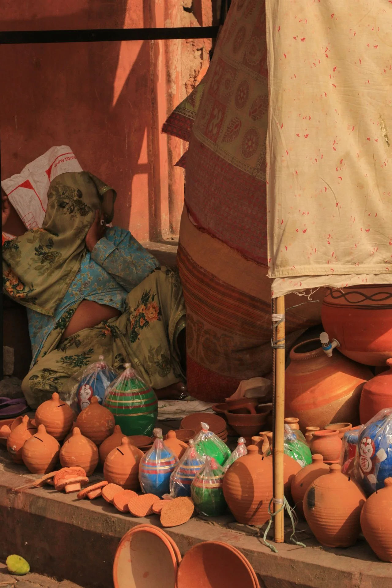 A woman resting on the ground among various clay pots and packaged items at a market stall. The scene is outdoors, with sunlight casting shadows.