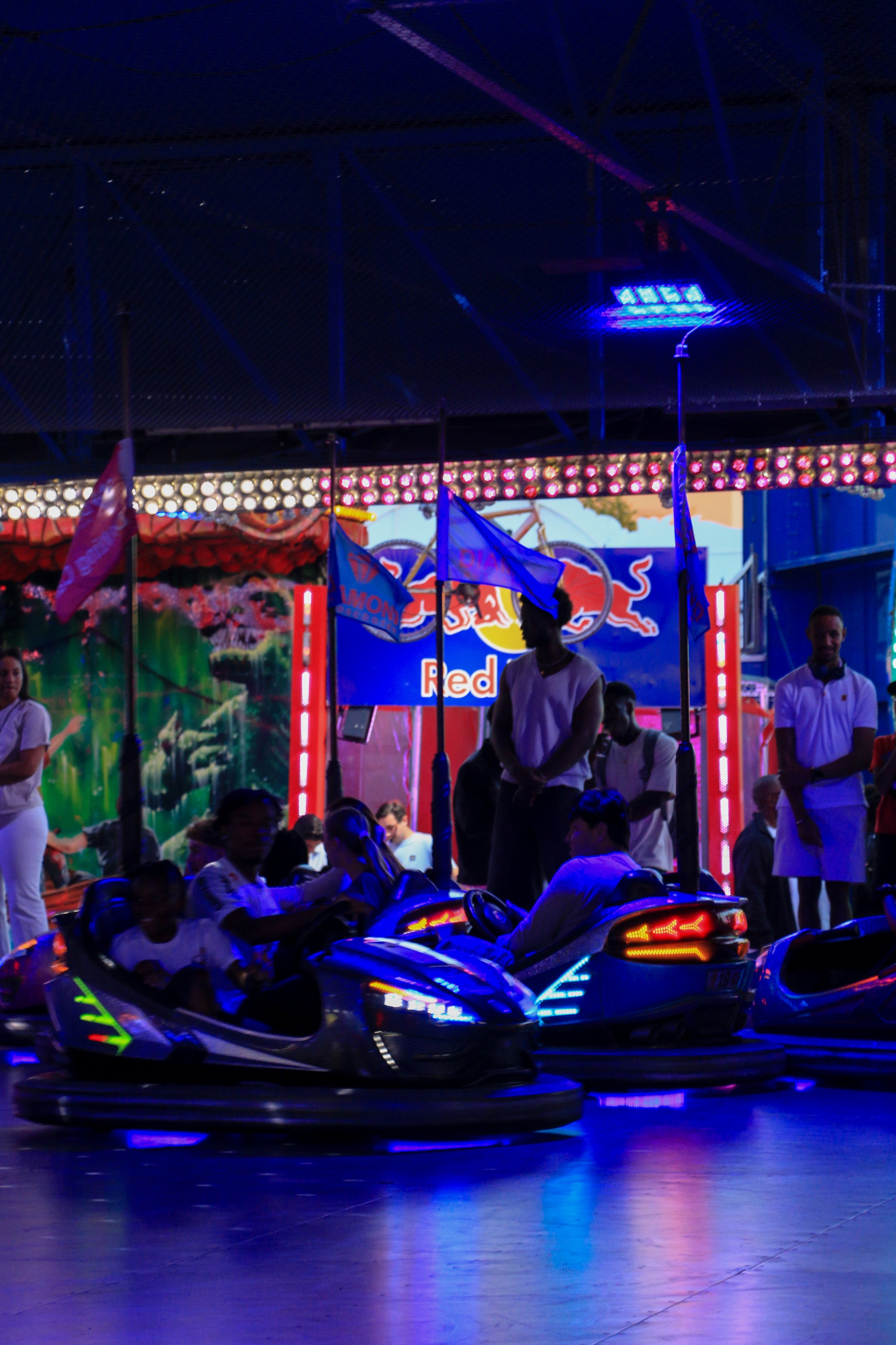 People riding bumper cars at an indoor amusement park with colorful lighting and a Red Bull banner in the background.