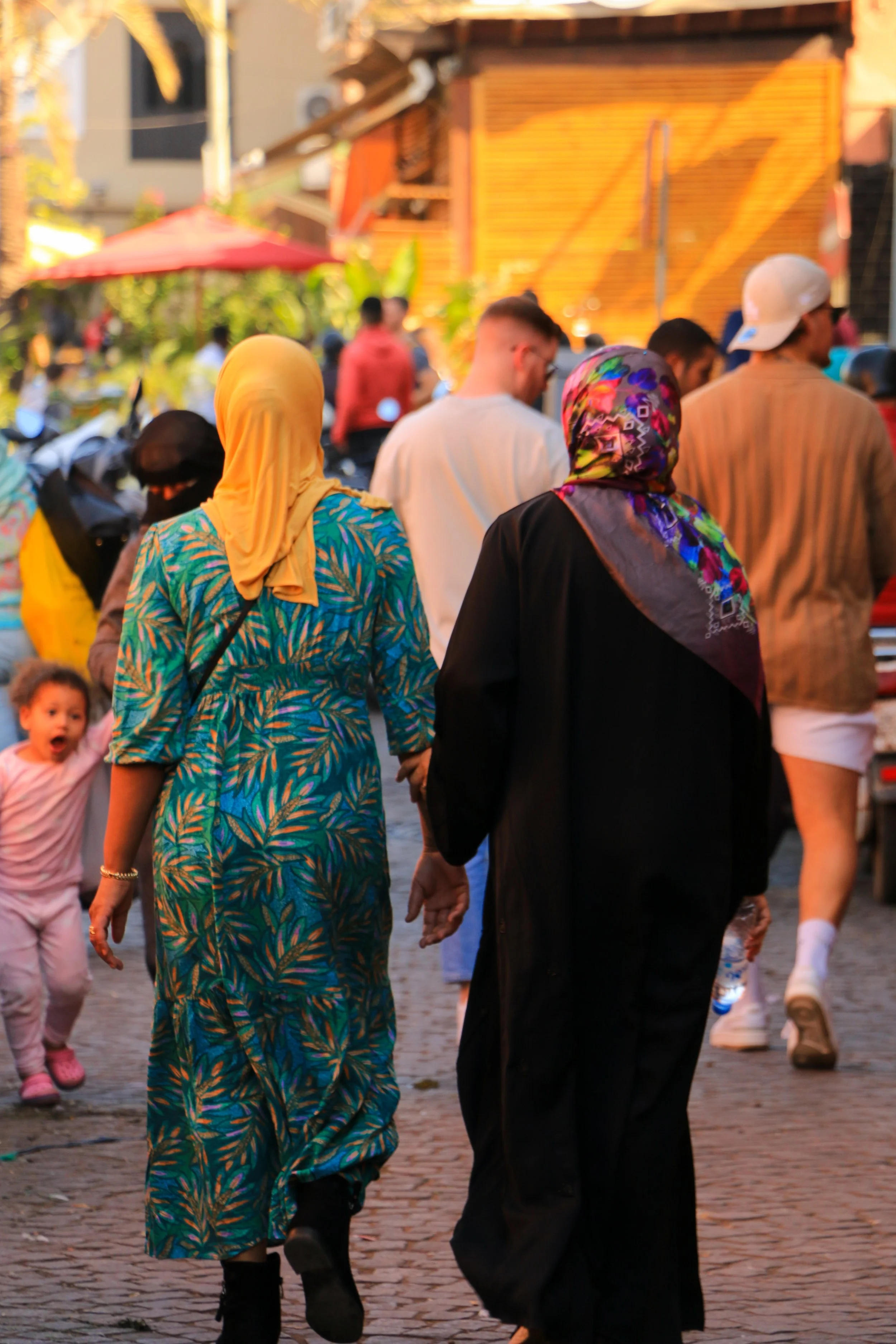 Two women, one in a yellow headscarf and colorful dress, and the other in a multicolored headscarf and black coat, hold hands as they walk on a busy outdoor street. Several people and a little girl are in the background.