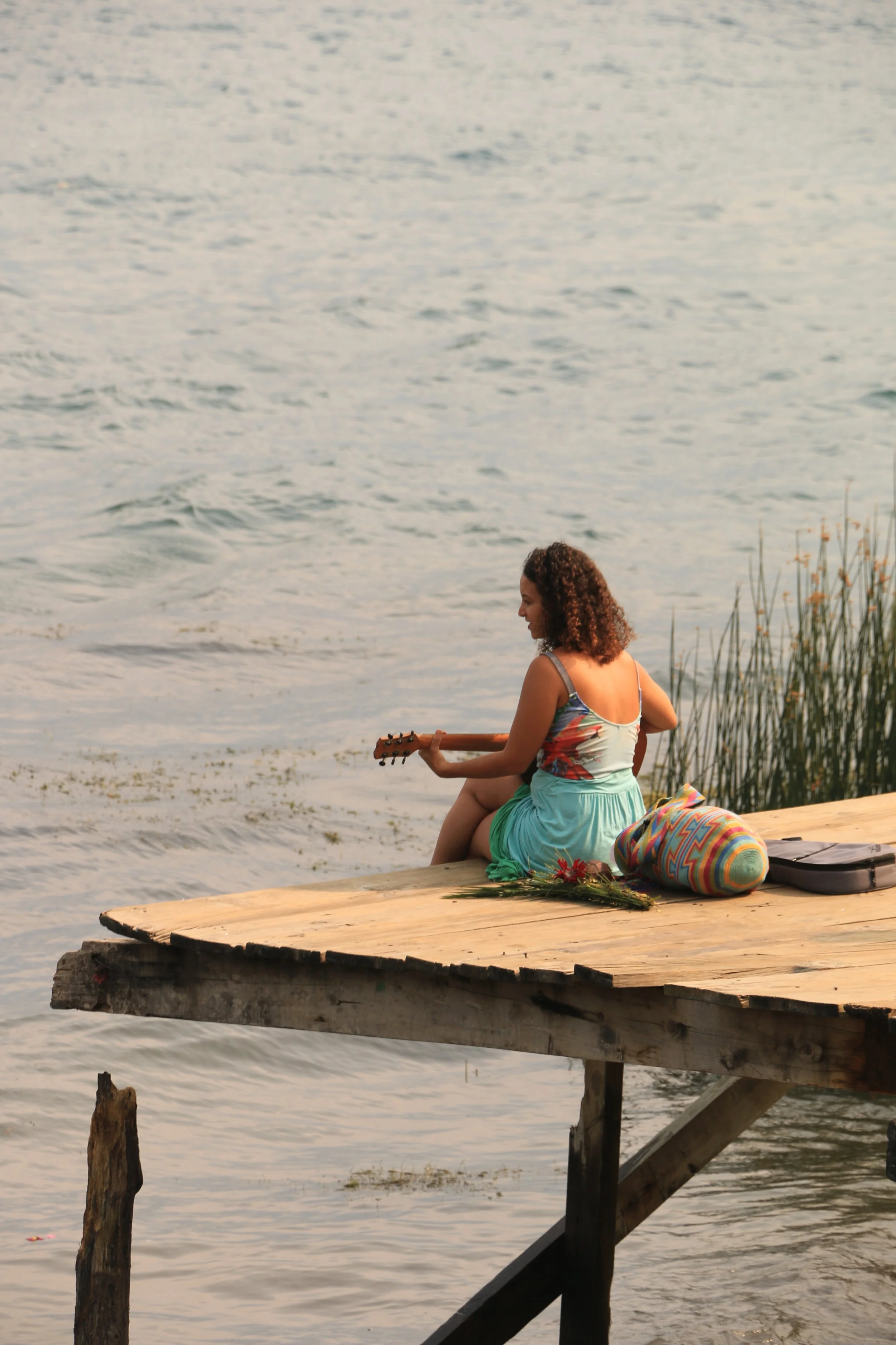 A woman with curly hair sitting on a wooden dock by the water, playing an acoustic guitar, with a colorful bag, flowers, and a black case beside her.