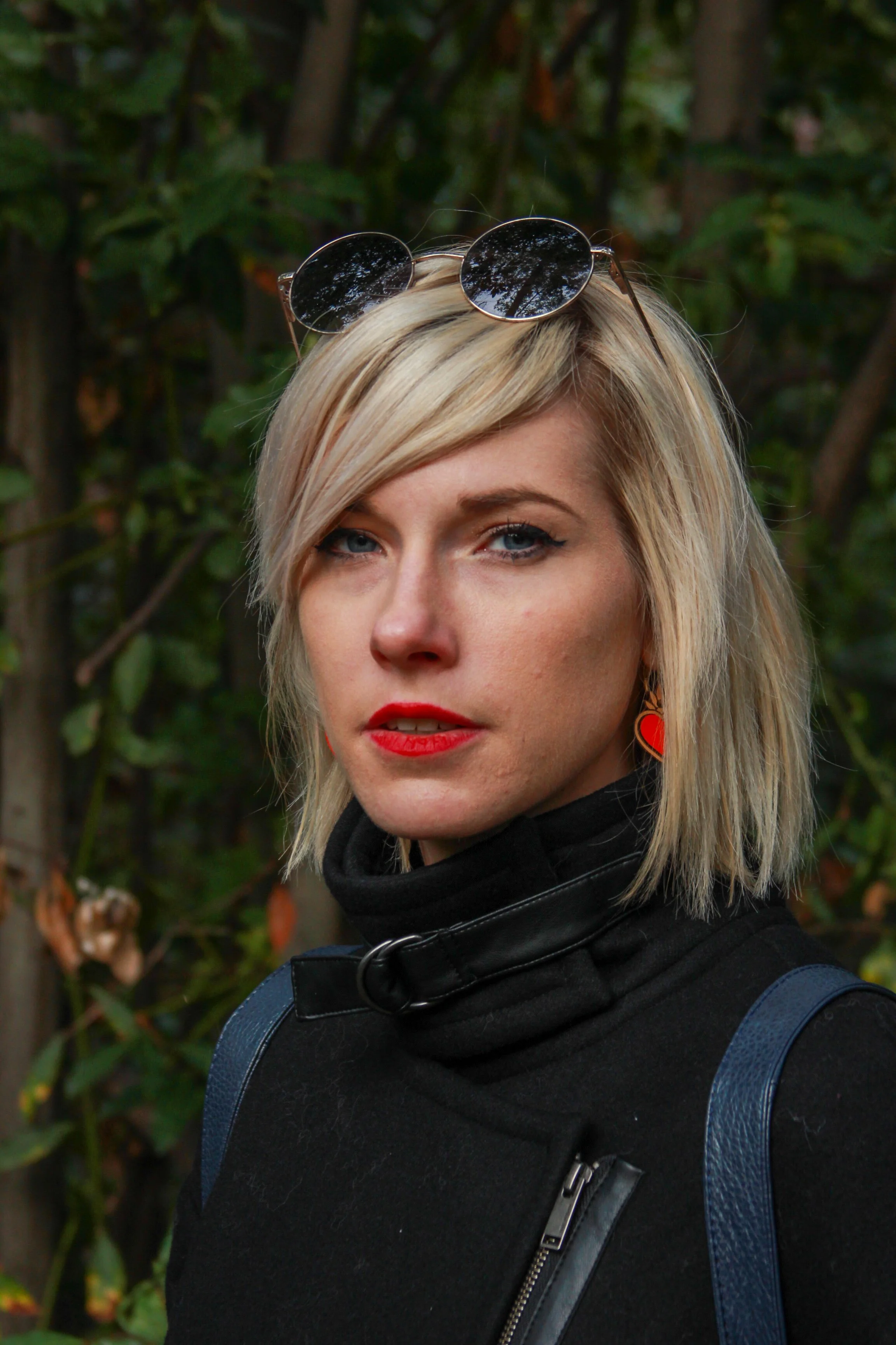 A woman with short blonde hair and blue eyes, wearing red lipstick, black jacket, and sunglasses on her head, standing outdoors amidst greenery.