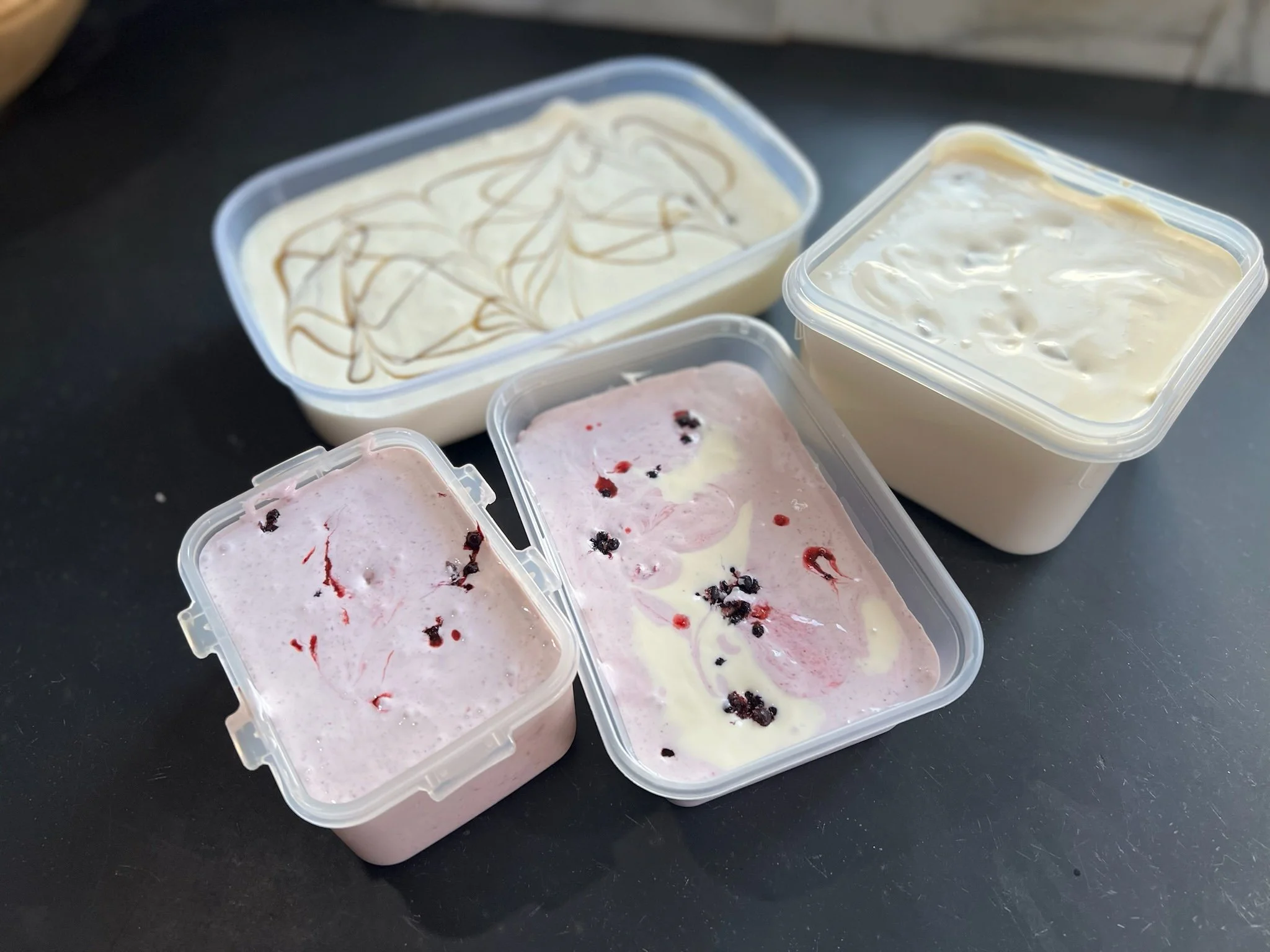 Various containers of homemade ice cream on a dark surface. The ice cream flavors include a pink berry swirl, vanilla with caramel drizzle, and plain vanilla. Berries are visible on some of the pink ice cream.