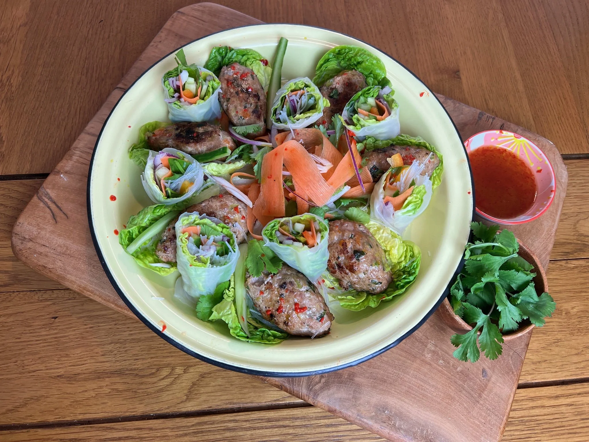 Plate of Vietnamese spring rolls with lettuce, grilled meat, and vegetables, served with a side of dipping sauce and fresh cilantro on a wooden board.