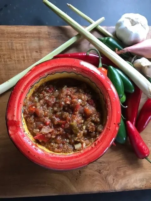 Traditional spicy sauce in a red pot surrounded by fresh lemongrass, chili peppers, and garlic on a wooden surface.