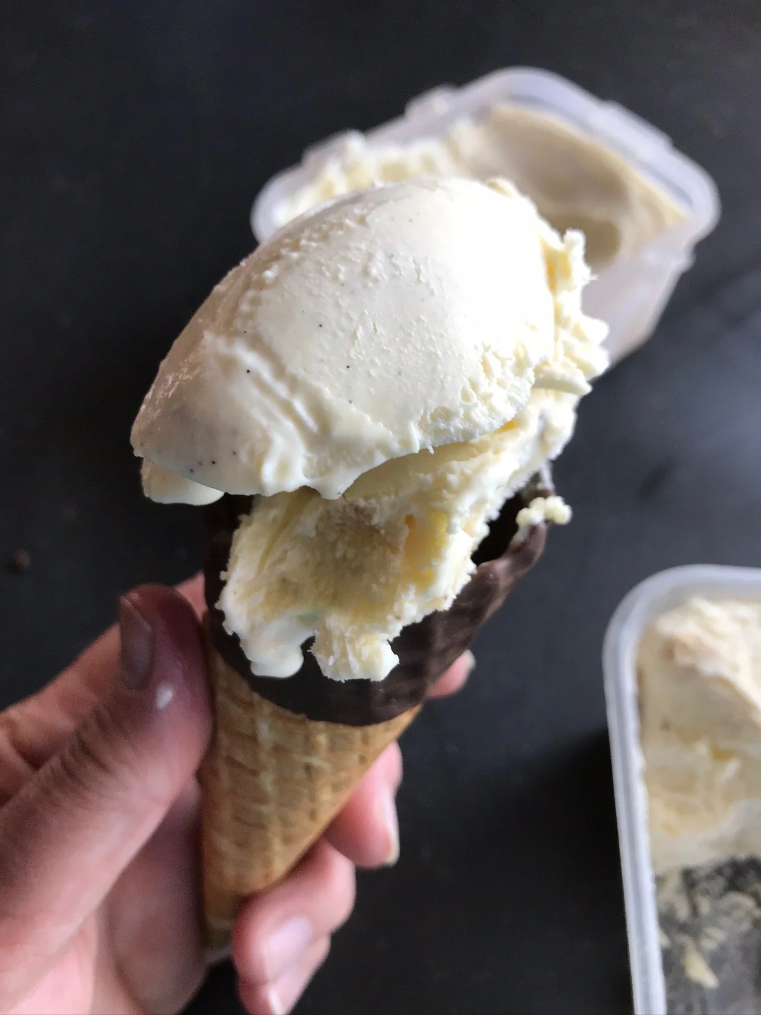 Ice cream cone with vanilla ice cream and chocolate-dipped cone held in a hand, with a container of ice cream in the background.