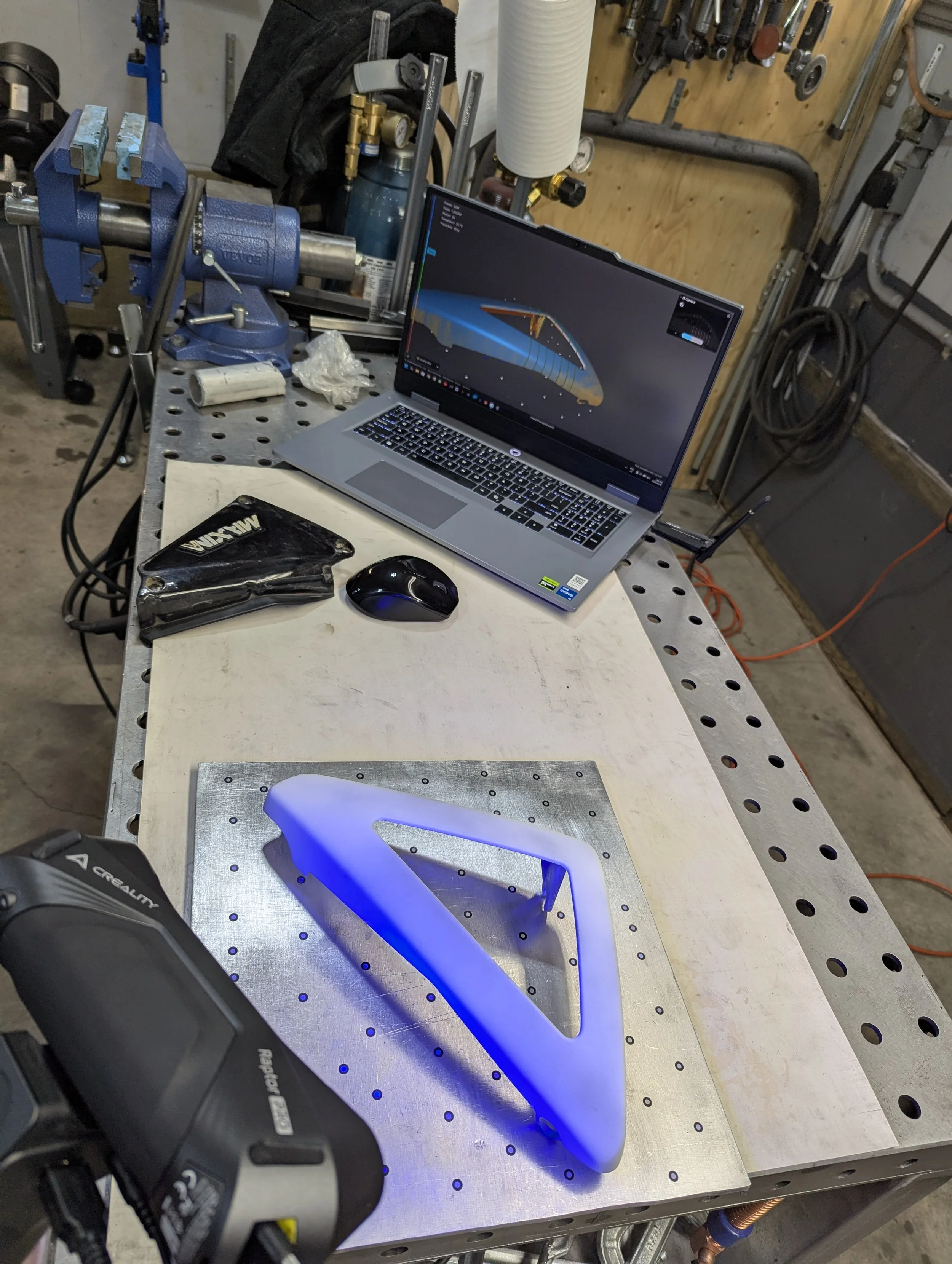 A workbench in a workshop with a laptop displaying a 3D model, a blue 3D printed part, a power tool, and various tools and equipment in the background.