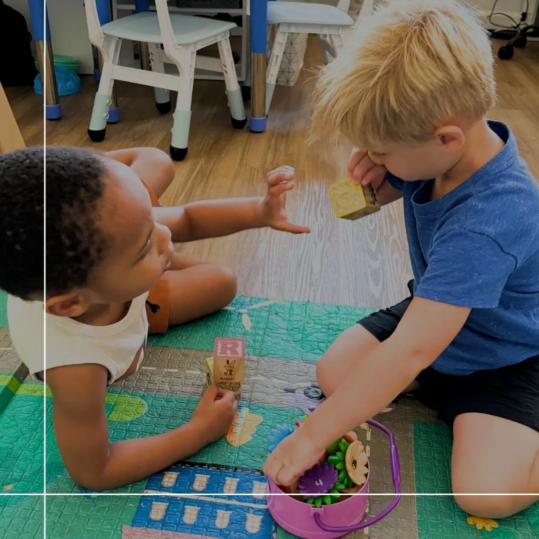Two young children sitting on the floor facing each other, engaging in a play activity with blocks. One child is holding a block with a letter 'R' on it. The scene takes place in a room with wooden flooring and some furniture in the background. Overlaid on the image is text that reads '5 Ways to Build Your Child's Social Confidence'.