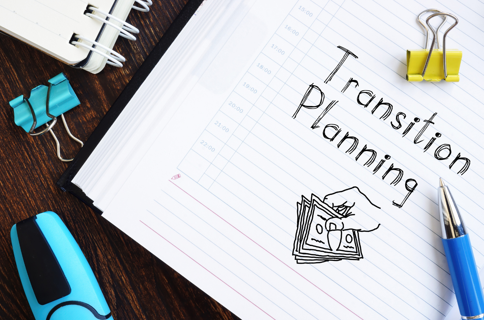 A notebook page with the words 'Transition Planning' written on it, surrounded by office supplies including binder clips, a pen, and a highlighter, on a wooden desk.