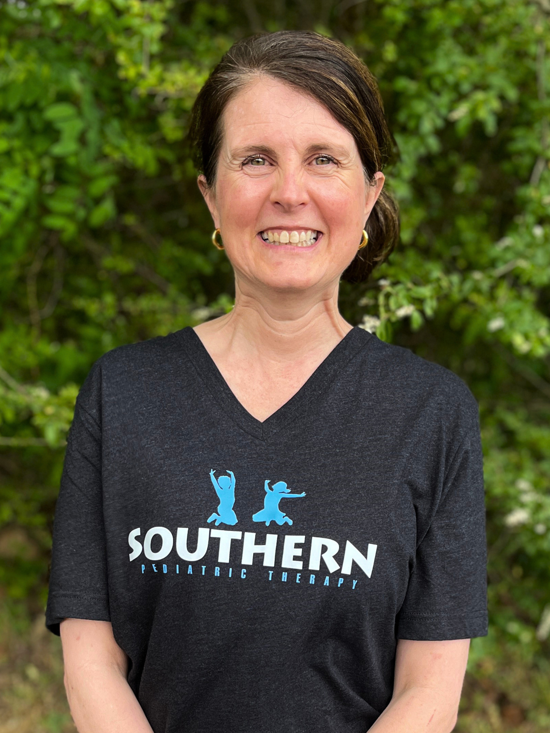 A woman with short dark hair and earrings smiling in front of greenery. She is wearing a black T-shirt with the words 'Southern Pediatric Therapy' and a logo of two jumping figures.