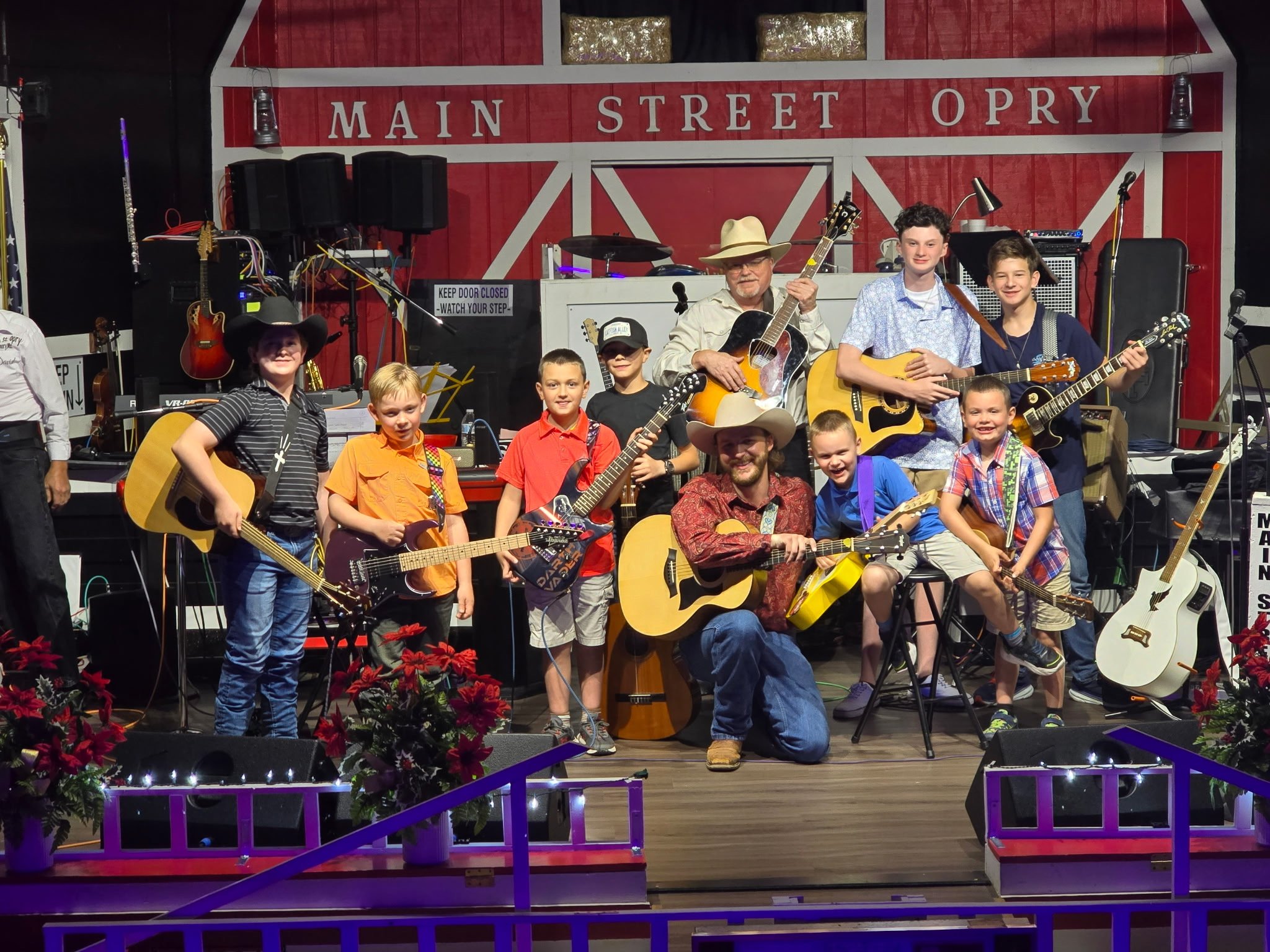 A group of children and adults with guitars on stage at the Main Street Opry, posing for a photo with a red and white backdrop. The stage is decorated with plants and lights.