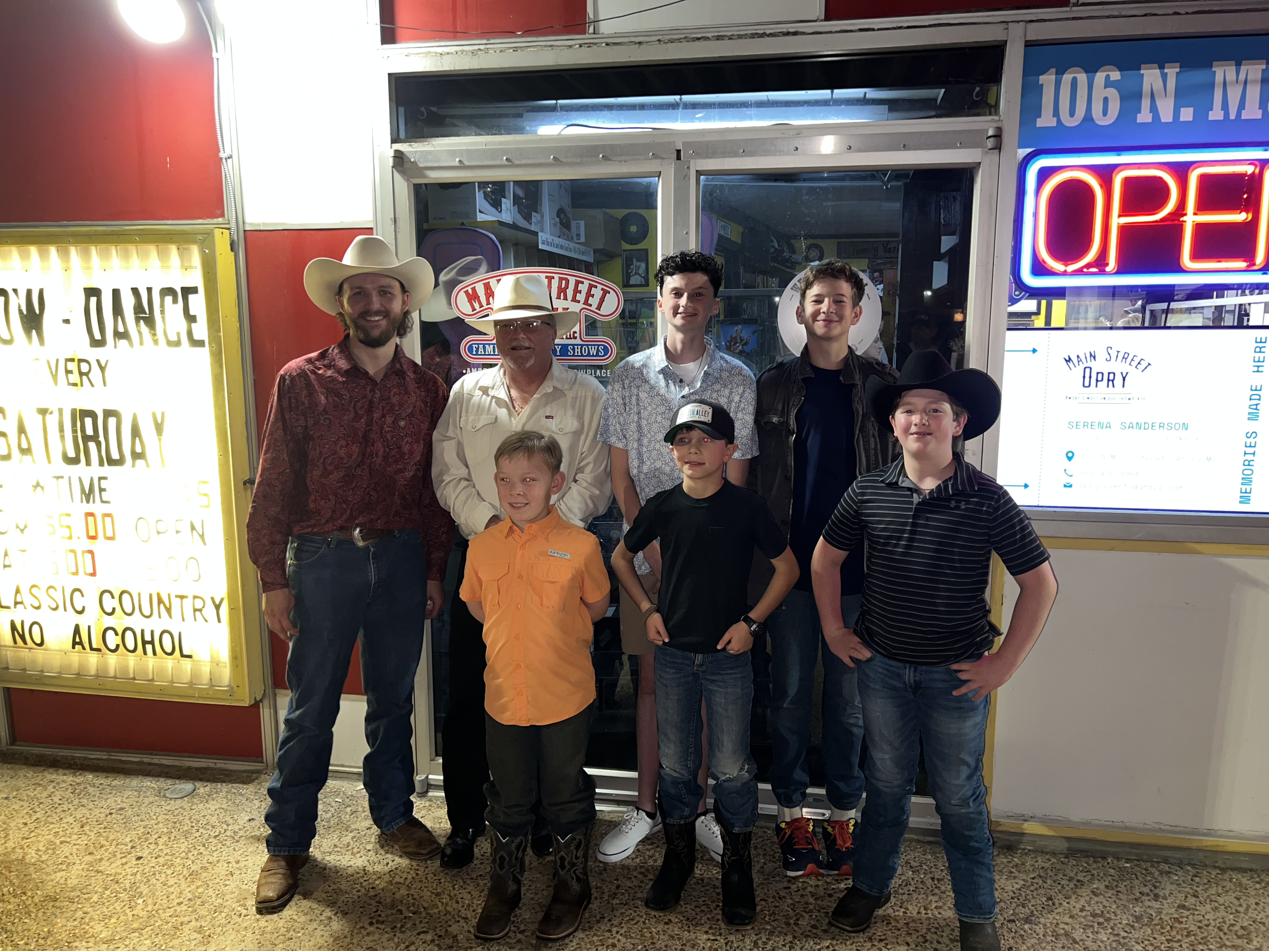 Group of seven people, including children and adults, posing outside a country-themed venue with neon signs and a yellow sign advertising a dance event.
