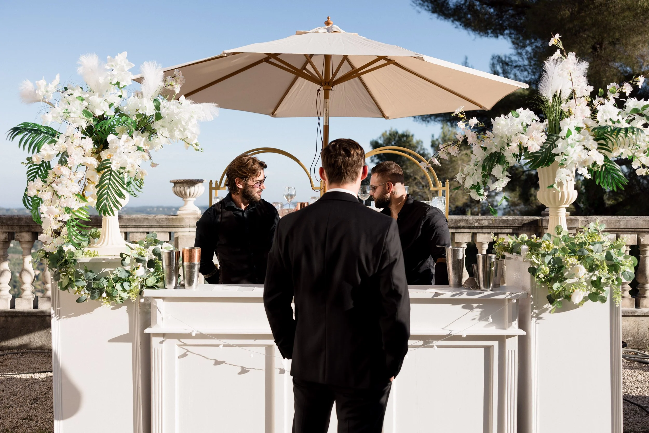 Trois hommes en costume noir discutent autour d'une table décorée de bouquets de fleurs blanches, sous un grand parasol beige en extérieur, avec un ciel bleu et des arbres en arrière-plan.