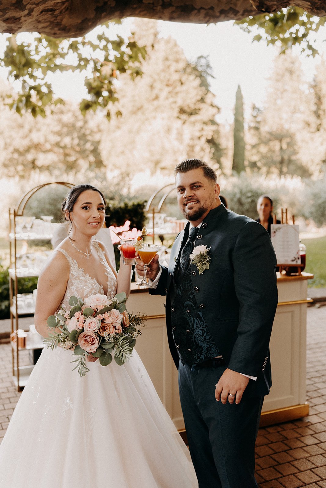 Un couple de mariés lors d'un mariage à l'extérieur, tenant des cocktails, entre eux. La mariée porte une robe de mariage blanche avec un bouquet de fleurs, et l'homme porte un costume noir avec une boutonnière. L'arrière-plan montre un décor de jardin avec des arbres en fleurs.