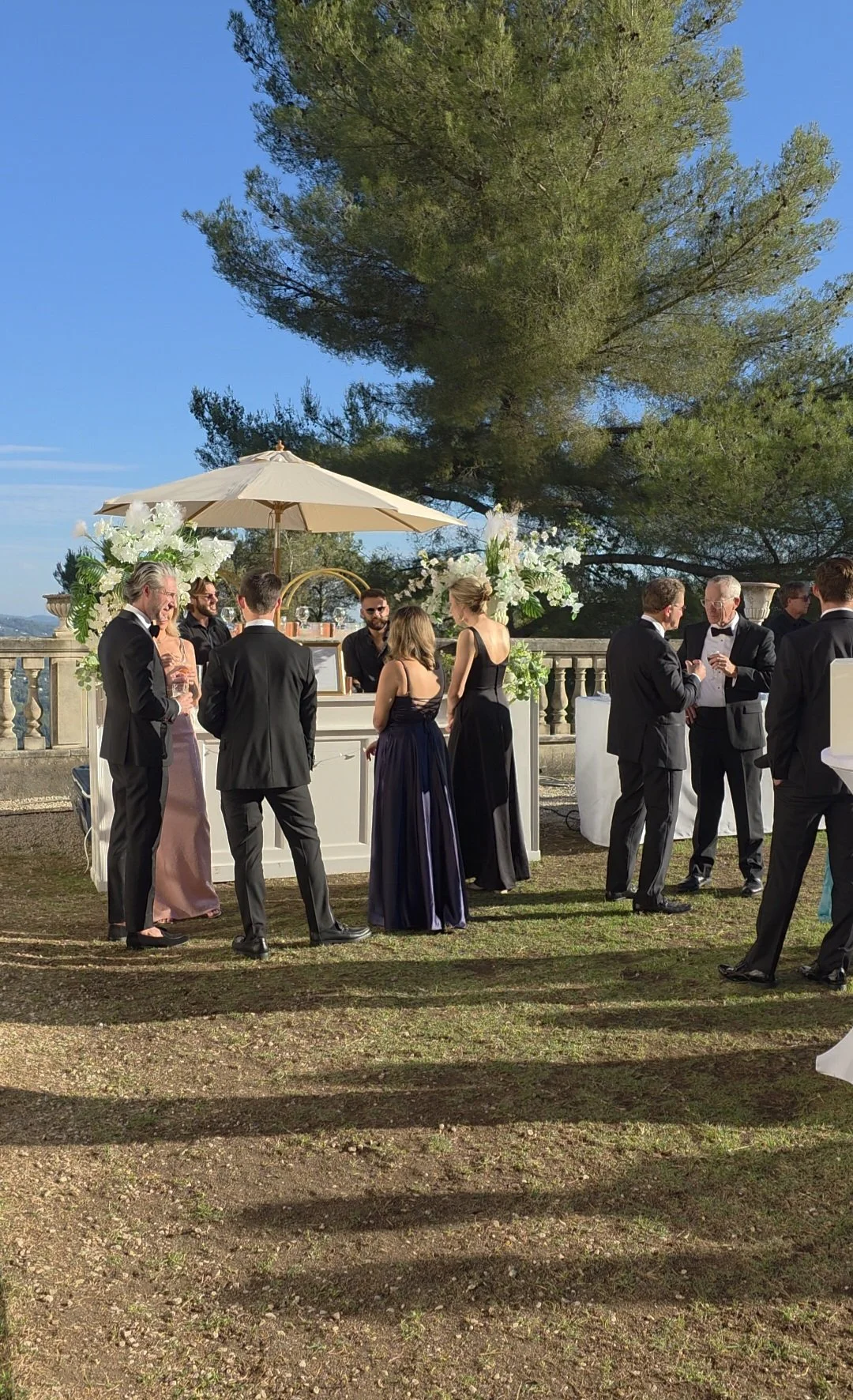 Groupe de personnes vêtues de vêtements formels lors d'un événement en extérieur, avec un bar sous un parasol et décorations florales, devant un grand arbre et un ciel bleu.