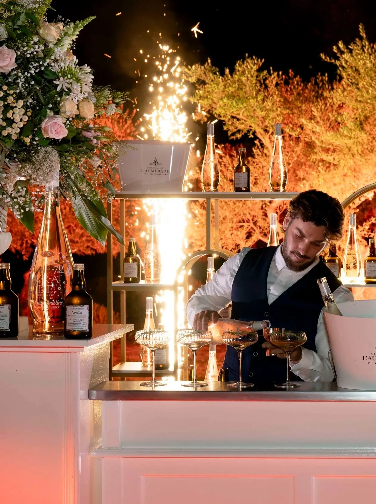 Un barman prépare des cocktails à un événement nocturne, avec des bouteilles de spiritueux, des fleurs et des feux d'artifice en arrière-plan.