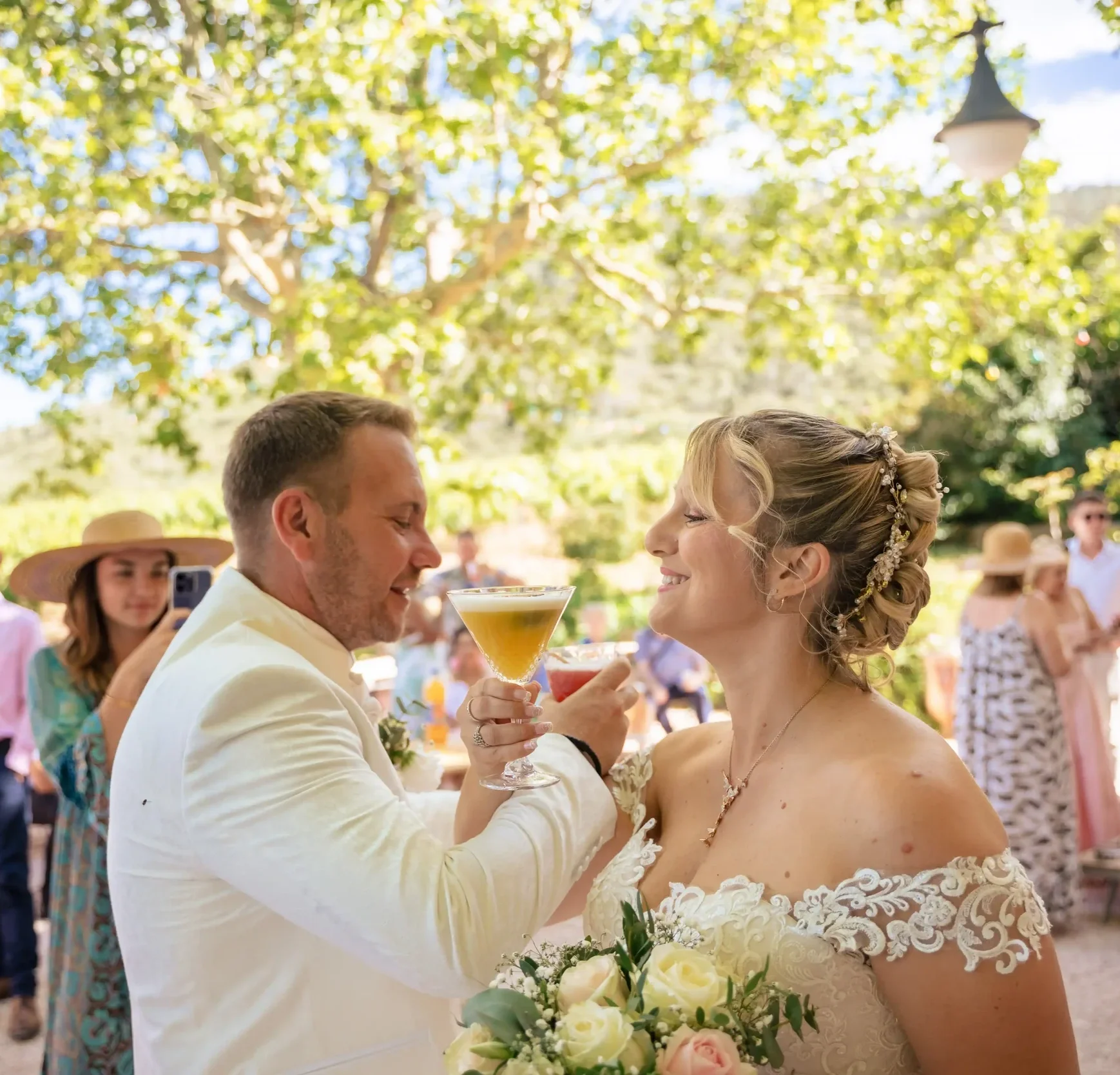 Un couple marié lors de leur mariage, s'embrassant avec des invités en arrière-plan dans un cadre extérieur avec des arbres et des invités vêtus de tenues estivales.