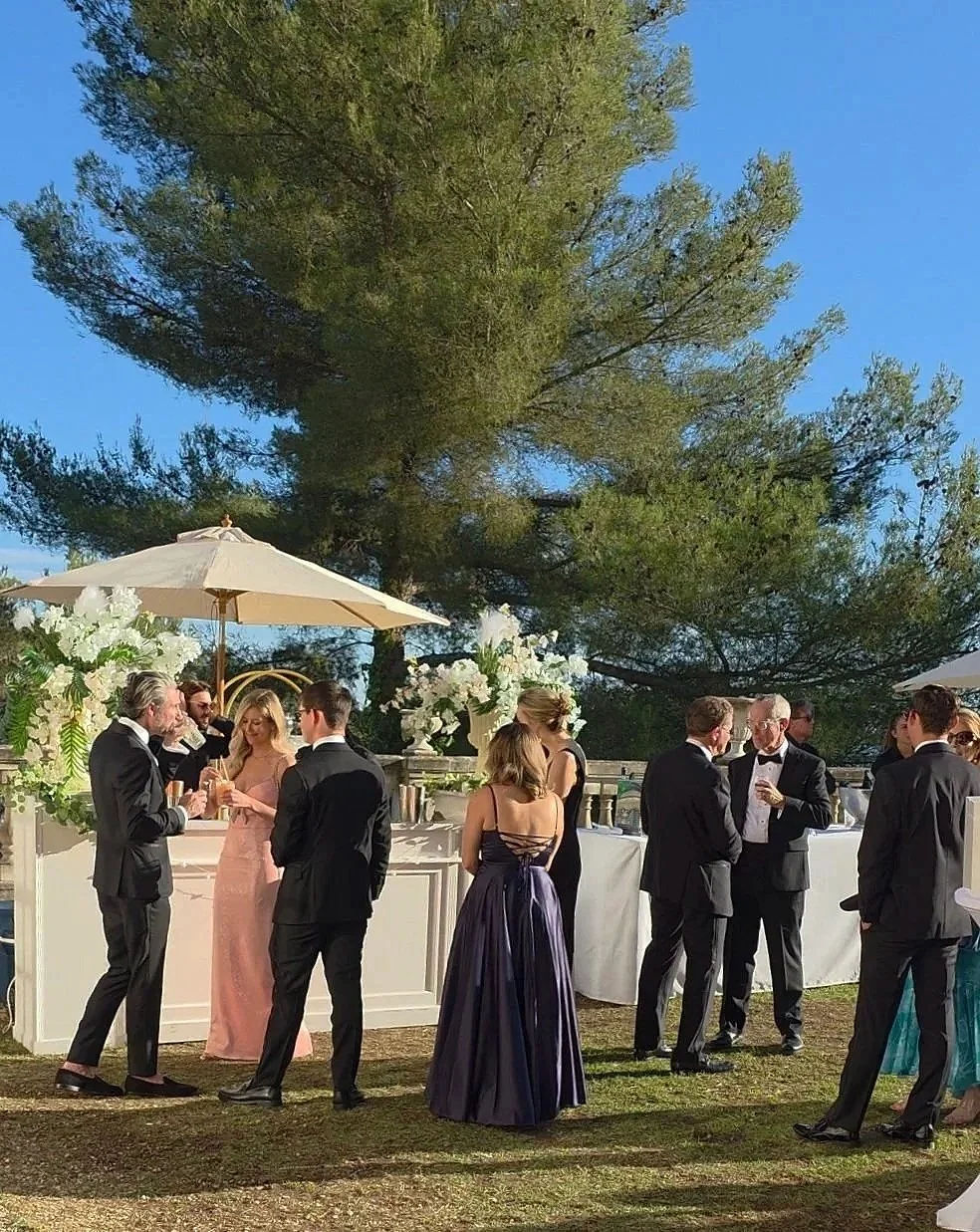 Groupe de personnes en tenues formelles lors d'une réception ou d'un cocktail en plein air sous un grand arbre, avec des décorations florales et des parasols.