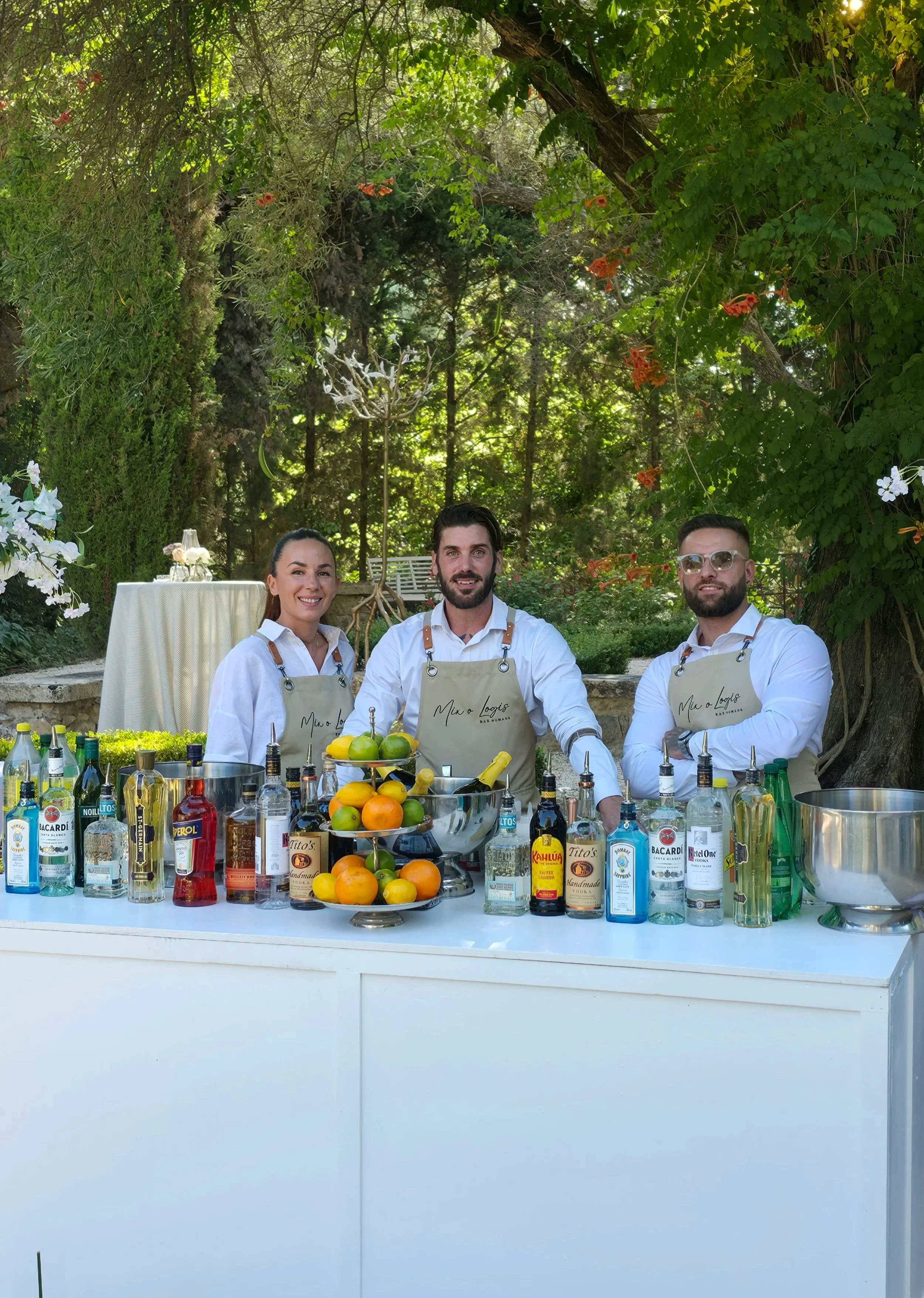 Trois personnes souriantes posant derrière un bar préparant des cocktails, avec un abondant assortiment de bouteilles d'alcool, de fruits frais comme des citrons, des limes et des oranges, ainsi que des éléments de décoration, en plein air dans un jardin avec des arbres et une végétation luxuriante.