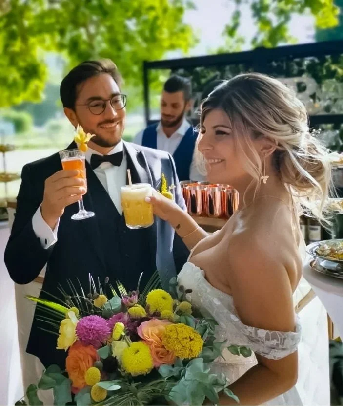 Un couple de mariés souriants lors d'une fête de mariage, échangeant des cocktails, la mariée tient un bouquet de fleurs colorées, dans un cadre extérieur ensoleillé.