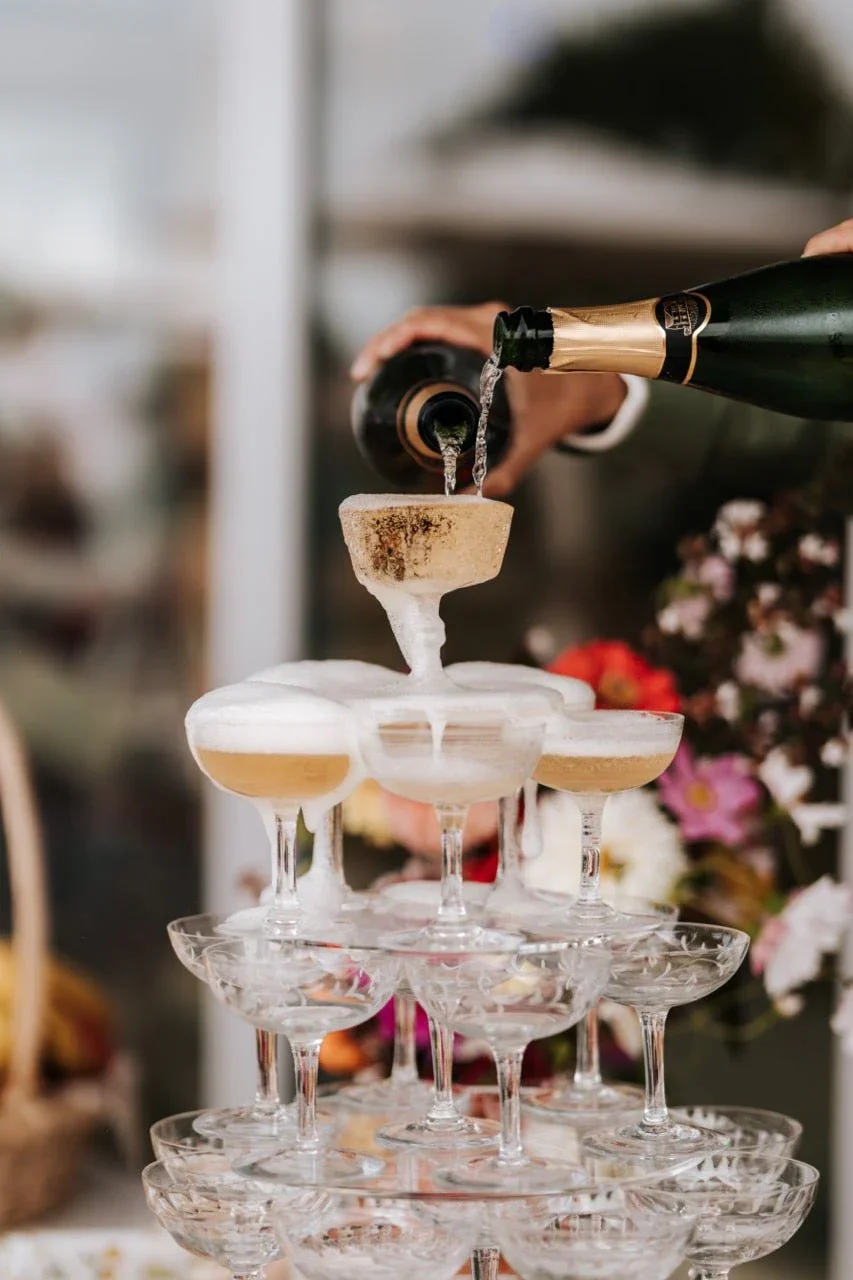 Une personne verse du champagne dans un pyramide de coupes à champagne, avec une décoration florale en arrière-plan.