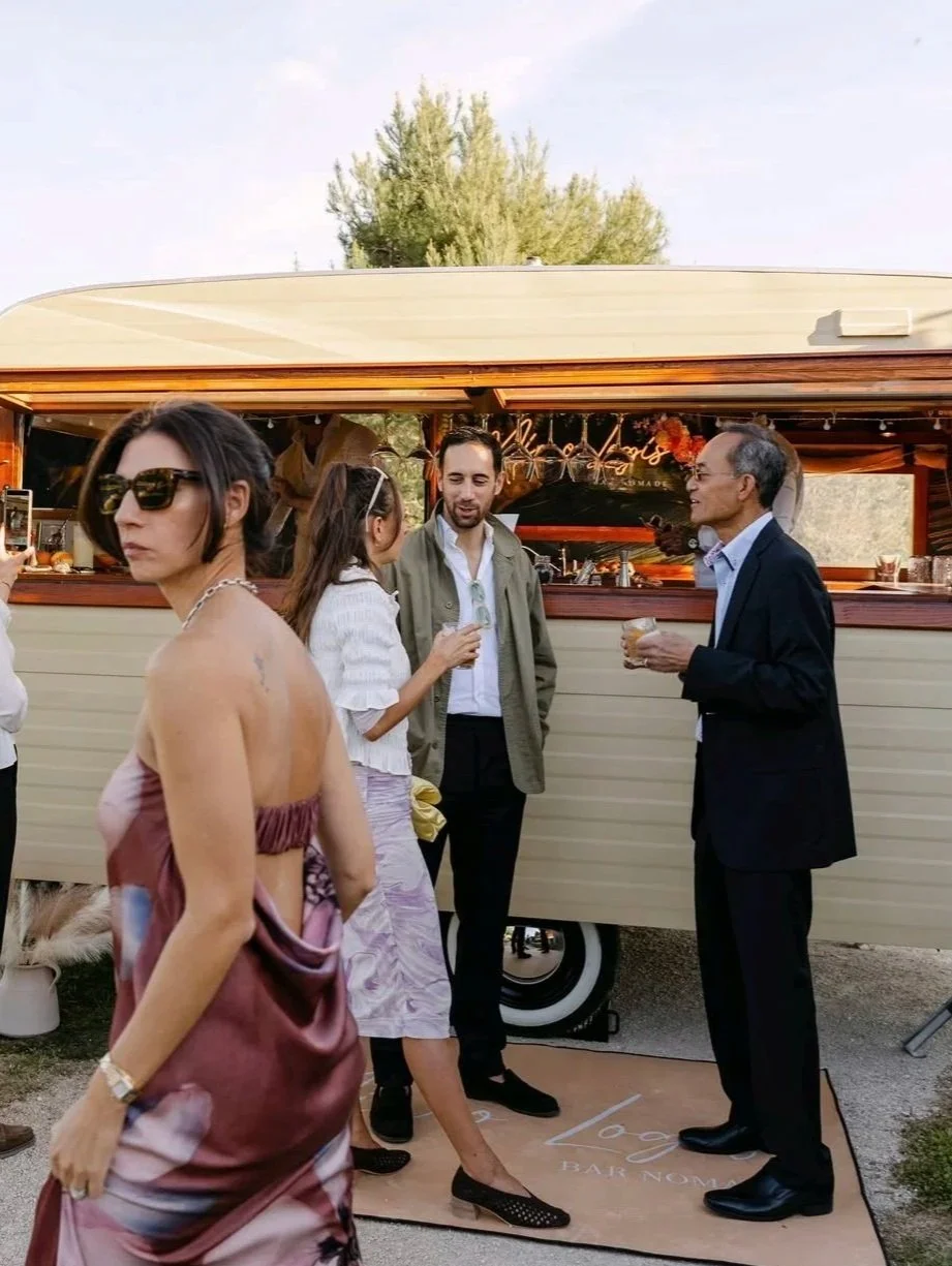 Groupe de personnes en conversation devant une roulotte bar avec arbres en arrière-plan, lors d'un événement extérieur en soirée.