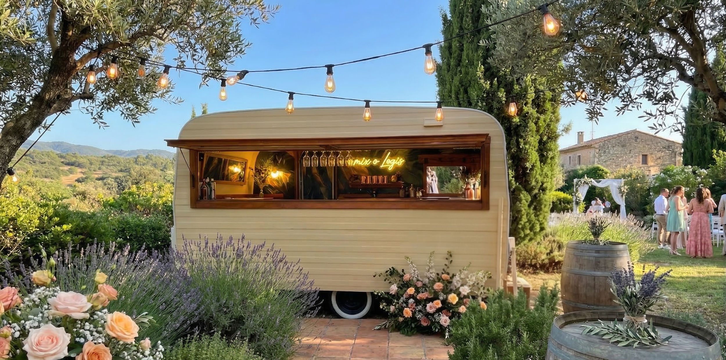 Un food truck blanc en forme de caravane dans un jardin fleuri, décoré de guirlandes lumineuses suspendues entre des arbres, avec des gens en arrière-plan lors d'une fête en plein air.