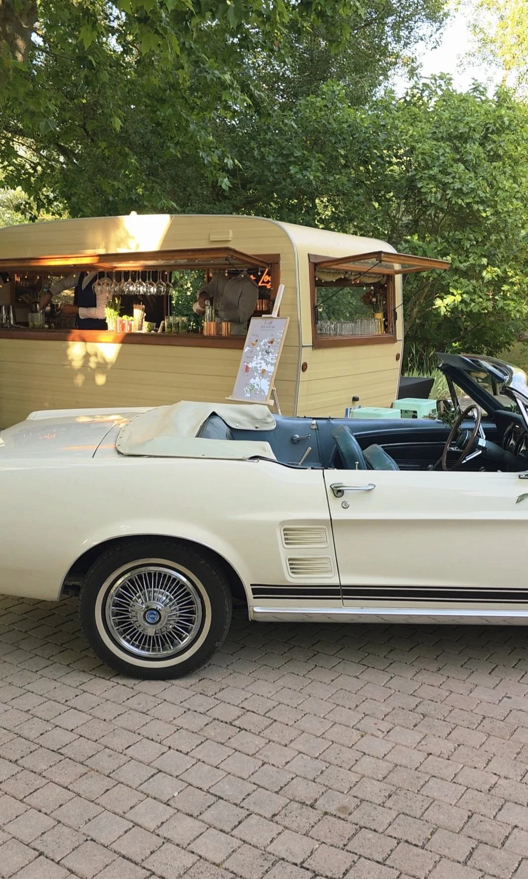 Voiture décapotable blanche avec intérieur bleu, stationnée sur un pavé, avec une roulotte ou un van vintage en arrière-plan, entourée d'arbres verts.