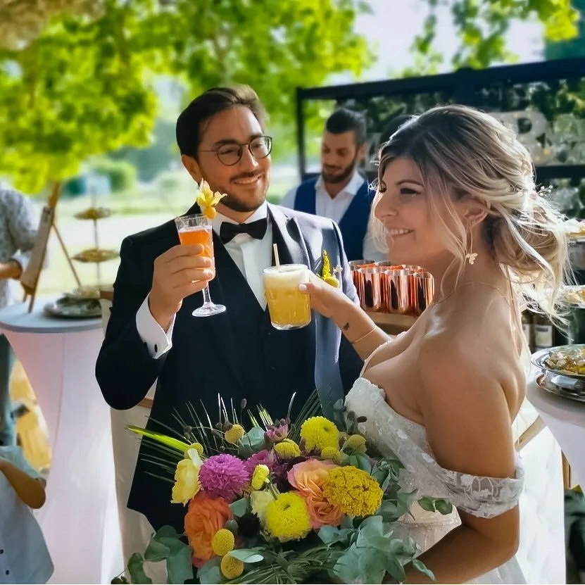 Un couple en costume de mariage souriant et levant des verres lors d'une réception en plein air, entourés d'invités et de décorations florales.