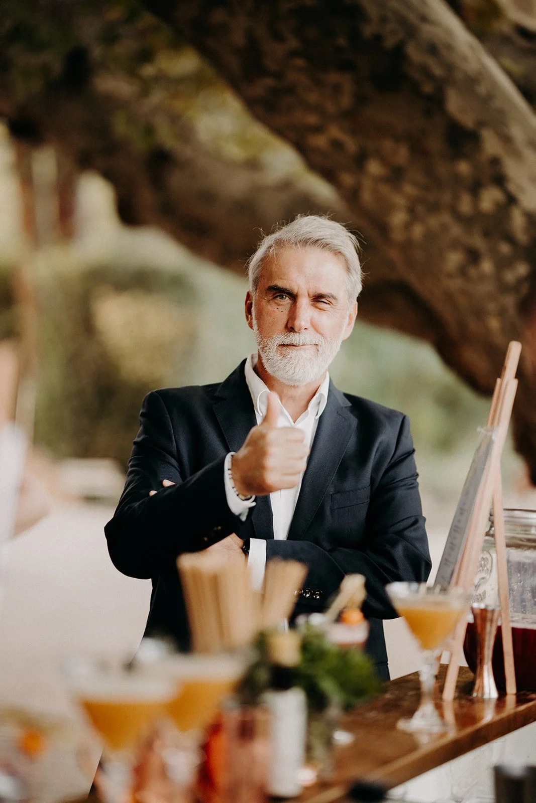 Homme mature en costume noir, souriant et faisant un geste de pouce levé, derrière un stand avec des boissons et des aliments dans un environnement naturel.