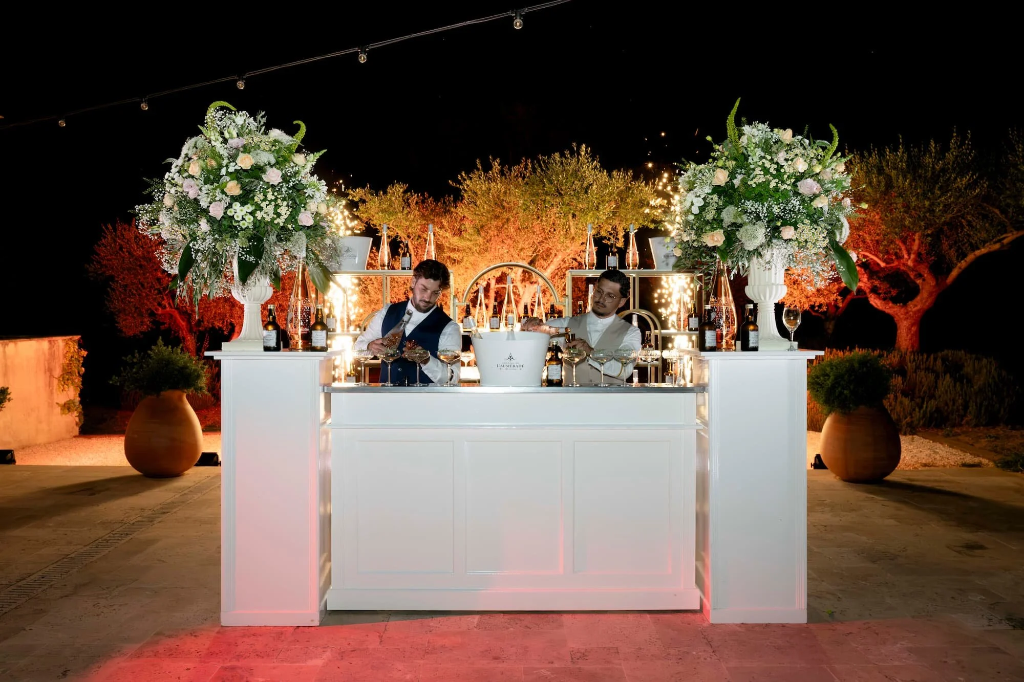 Deux barmans servent des cocktails derrière un bar blanc décoré de grandes compositions florales, lors d’une réception nocturne en plein air.