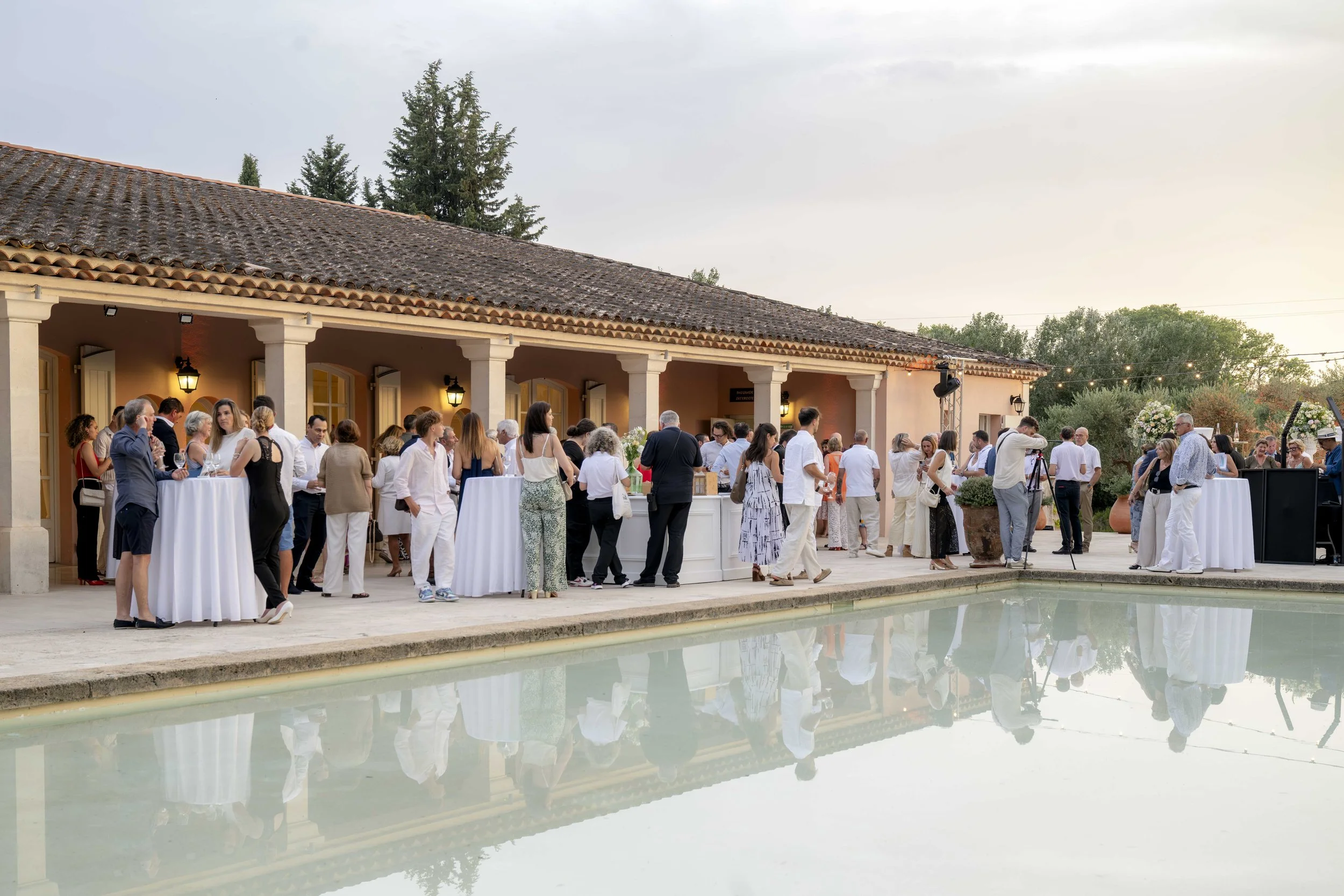 Groupe de personnes à une fête ou un événement en plein air à côté d'une piscine, sous un bâtiment avec un toit en tuiles. Certaines personnes discutent, d'autres regardent vers la scène où un DJ joue de la musique.
