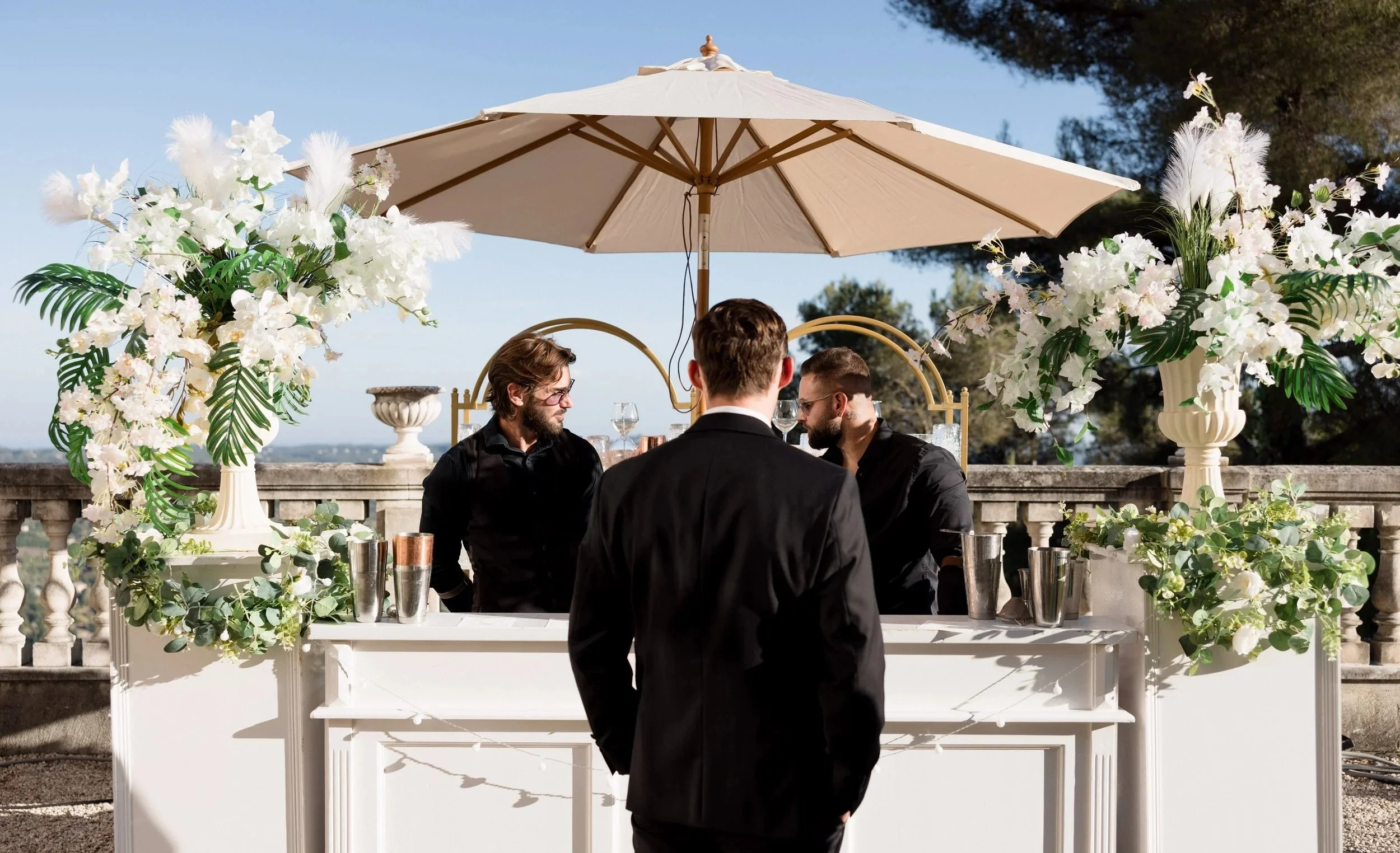 Trois hommes habillés en noir se tiennent devant un bar à cocktail mobile décorée de fleurs blanches et de plantes vertes, avec un grand parasol beige au-dessus, dans un cadre extérieur ensoleillé.
