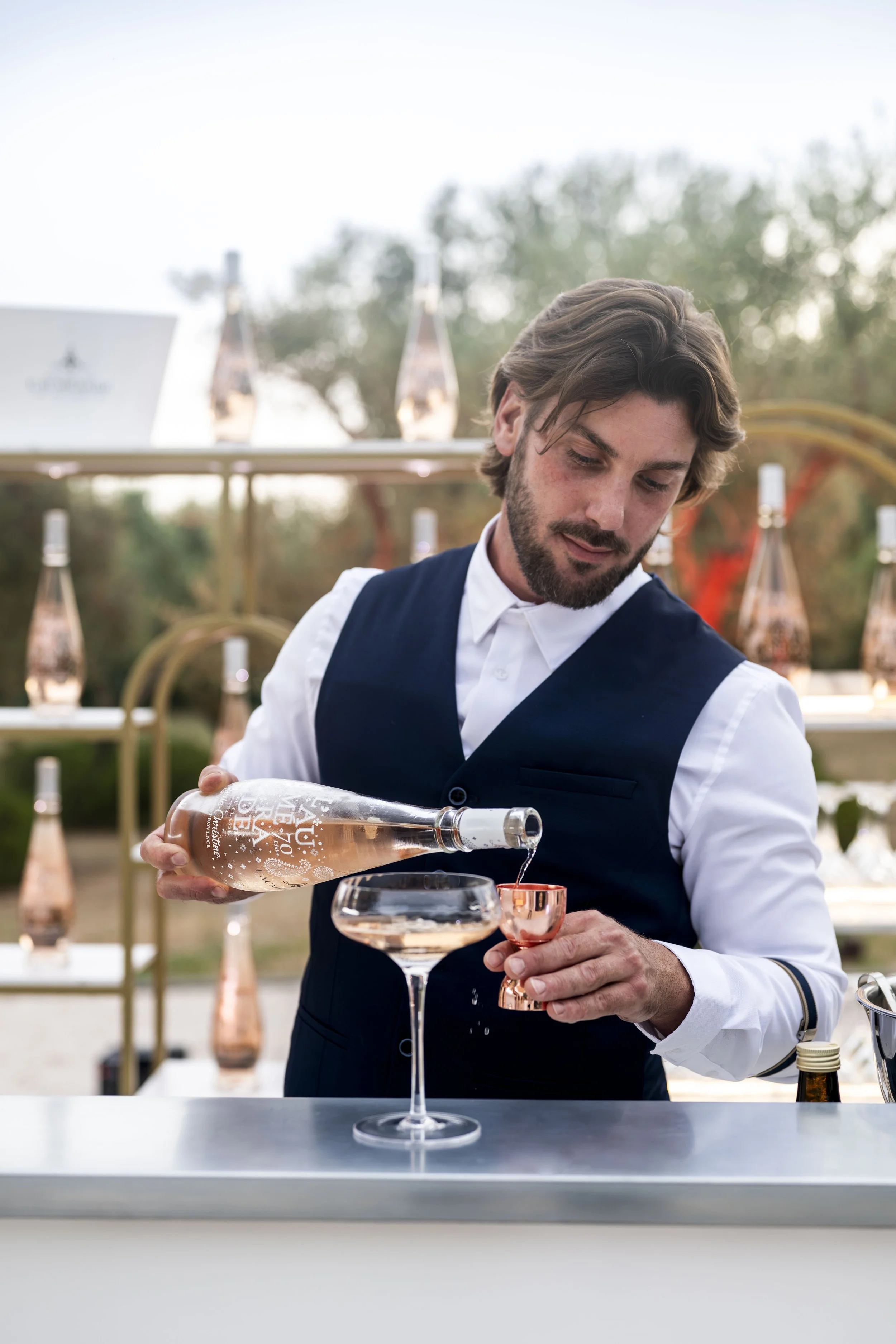 Un serveur en costume verse une boisson rosée dans un verre à cocktail en acier cuivré lors d'un événement en plein air.