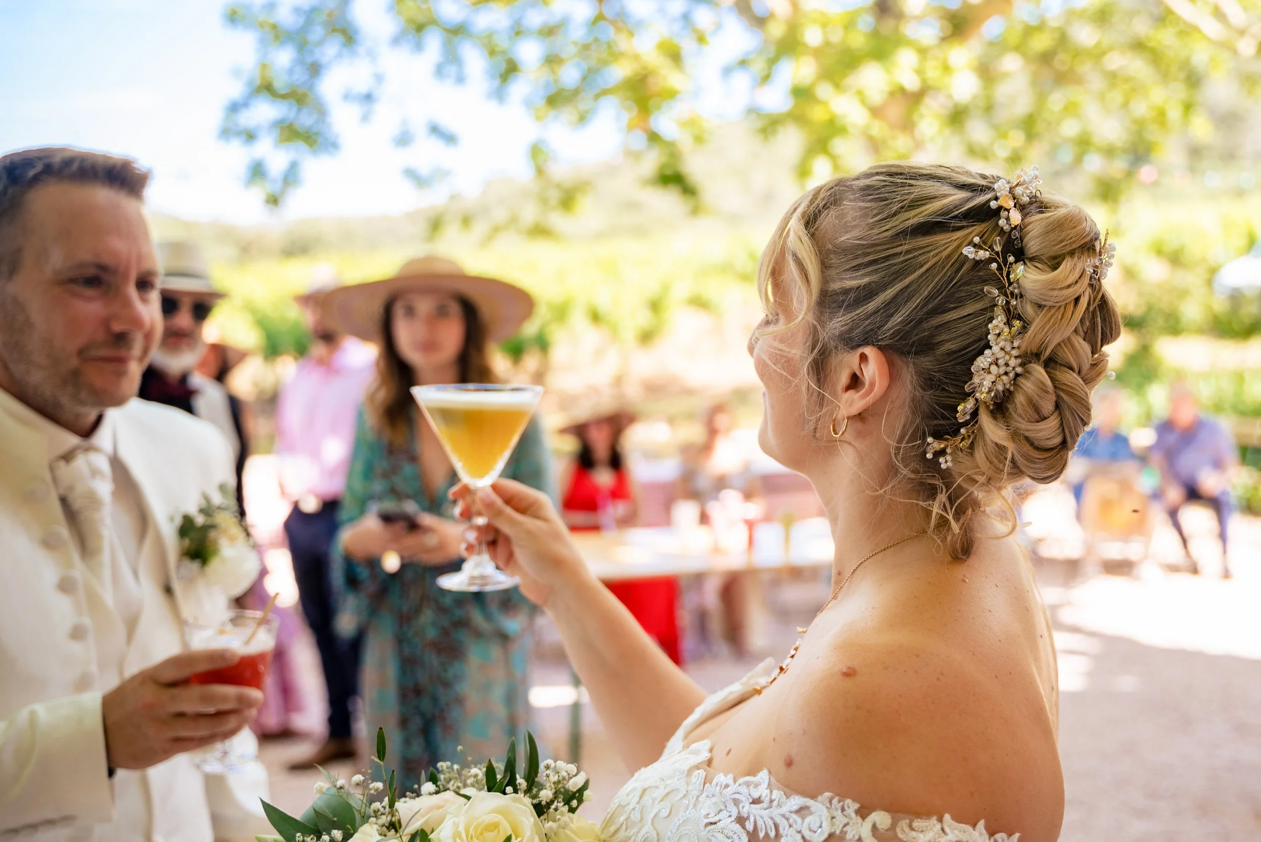 Une mariée tenant un verre à cocktail lors d'un mariage en extérieur, entourée d'invités en arrière-plan.