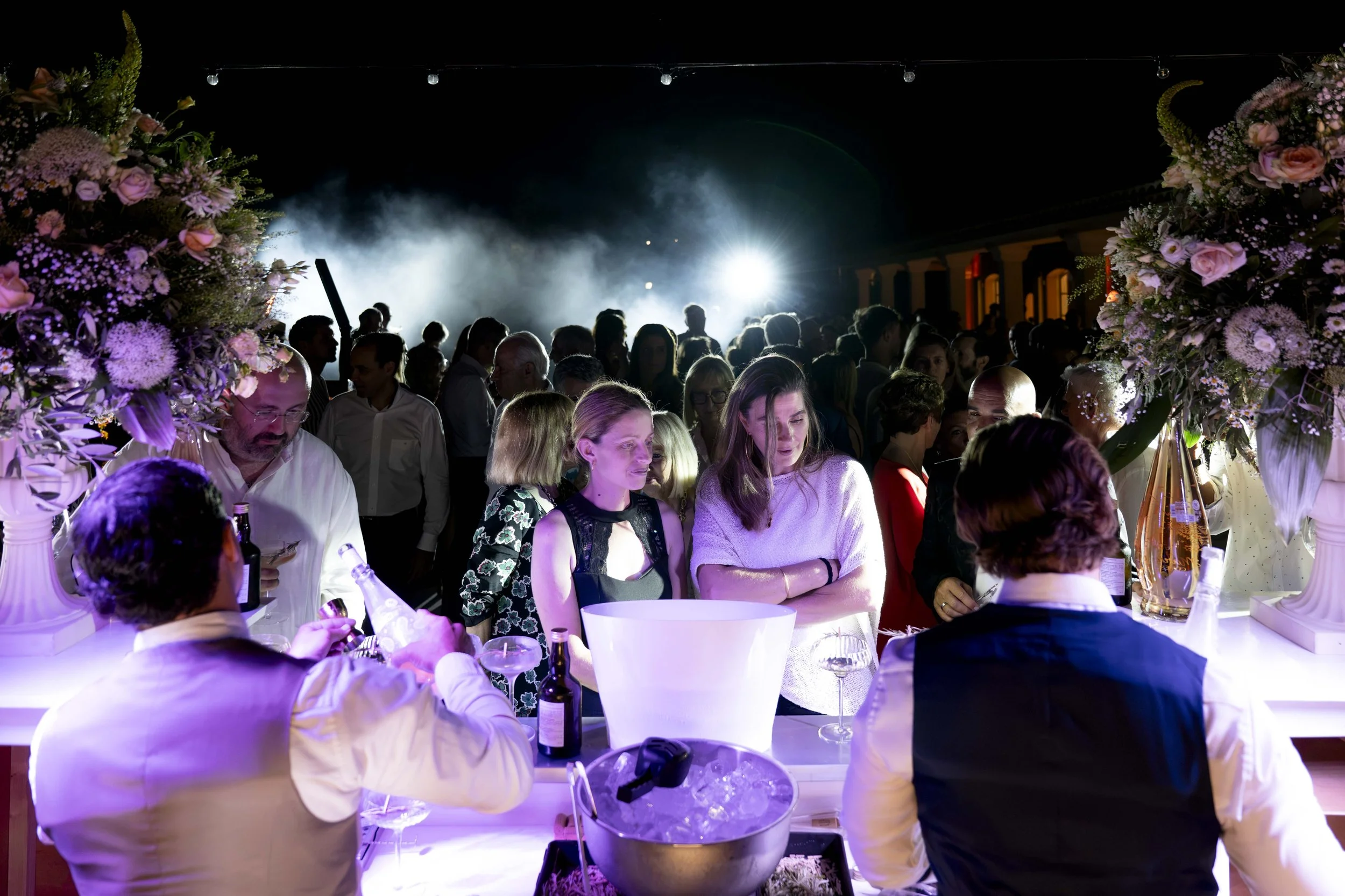 Une scène de fête ou de réception avec de nombreux invités, décorée avec de grandes compositions florales et éclairages lumineux.