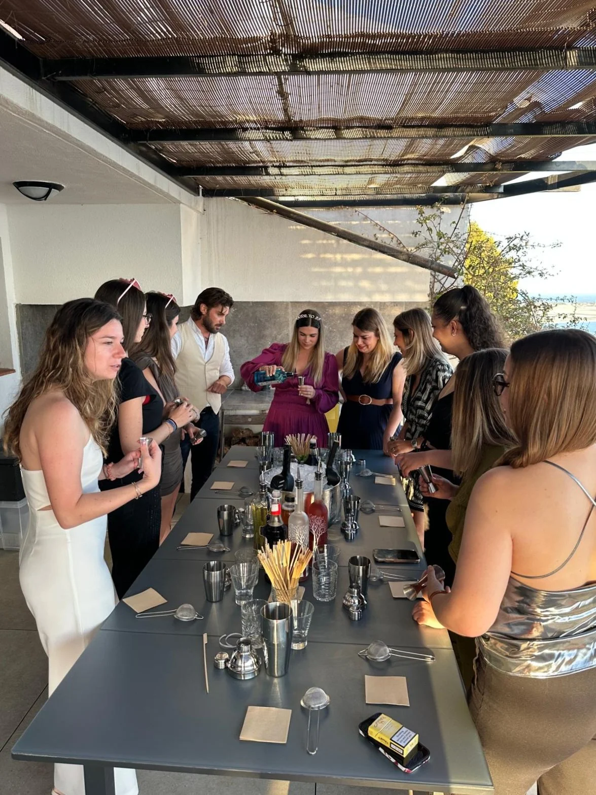 Groupe de personnes lors d'une dégustation de vin ou d'une activité sociale, autour d'une grande table avec des verres, bouteilles et accessoires de bar, dans un espace intérieur ou semi-ouvert.