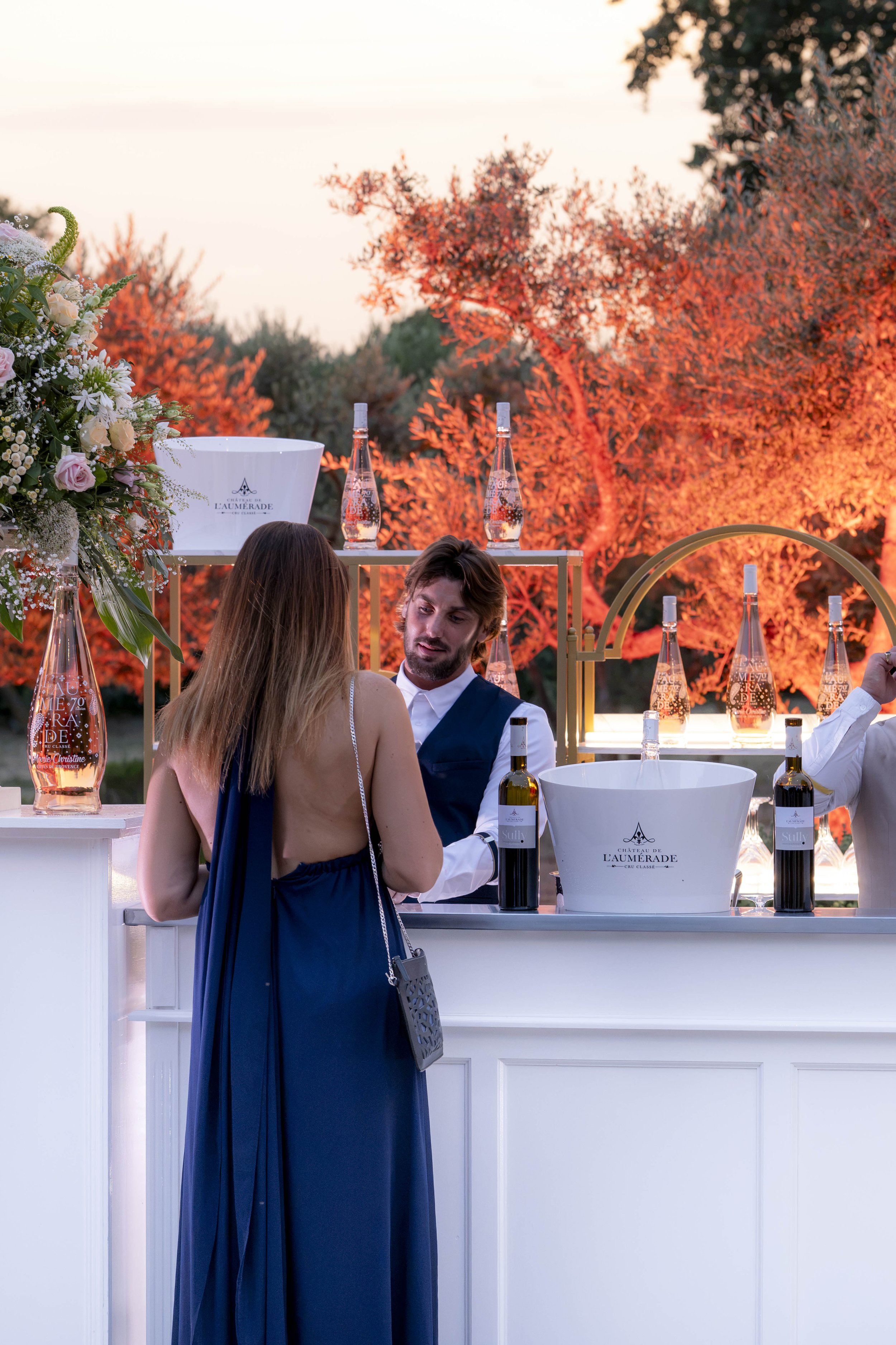 Une scène élégante en plein air avec une femme en robe bleue et un homme en veste blanche, échangeant des mots devant un bar avec des bouteilles de vin et des bouteilles d'eau Titanic sur un comptoir blanc. En arrière-plan, des arbres illuminés en rouge créent une ambiance chaleureuse et sophistiquée lors d'une soirée.