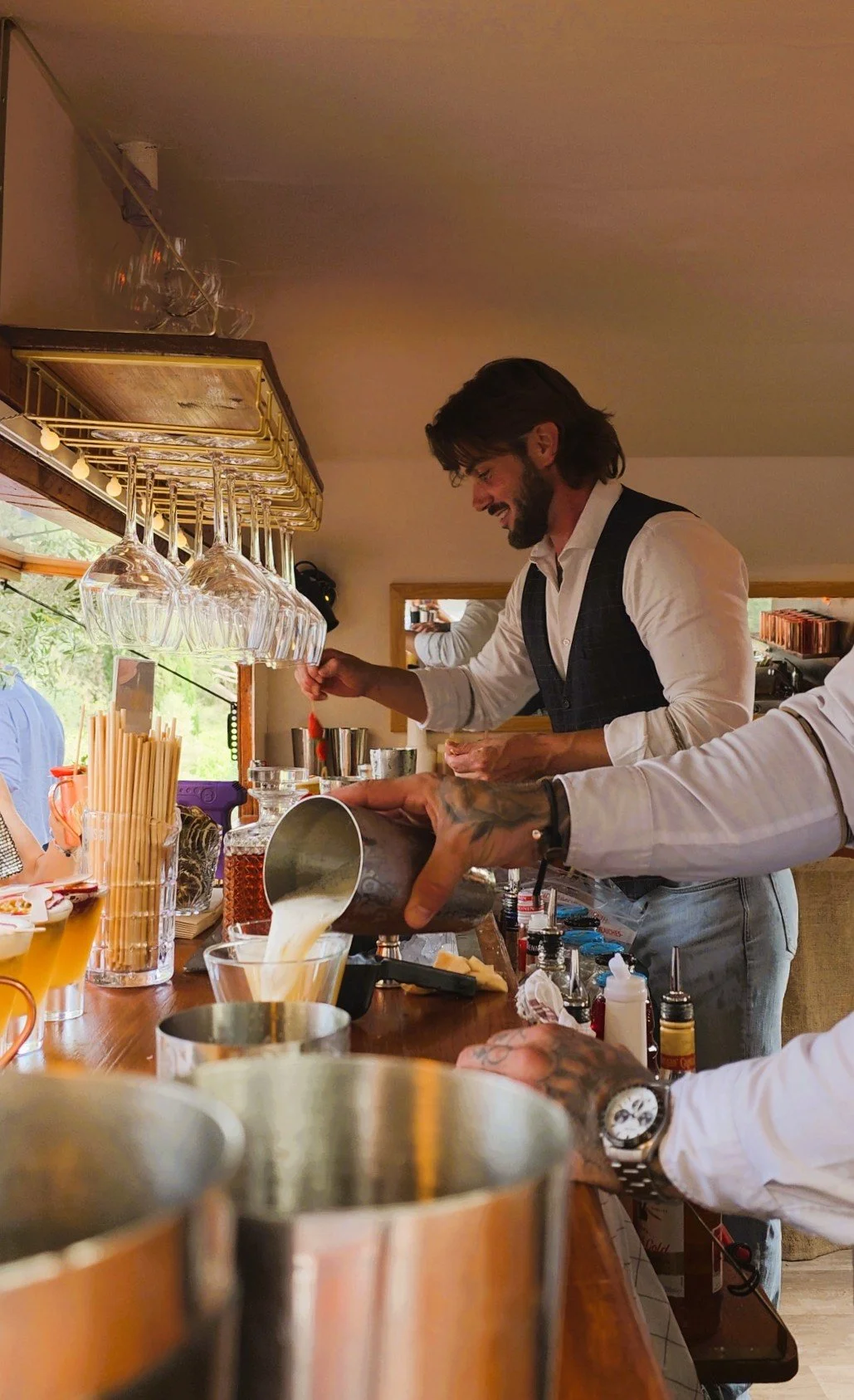 Deux hommes préparent des cocktails dans un bar, avec des verres à vin suspendus au-dessus du bar, un bar en bois, et une ambiance chaleureuse.