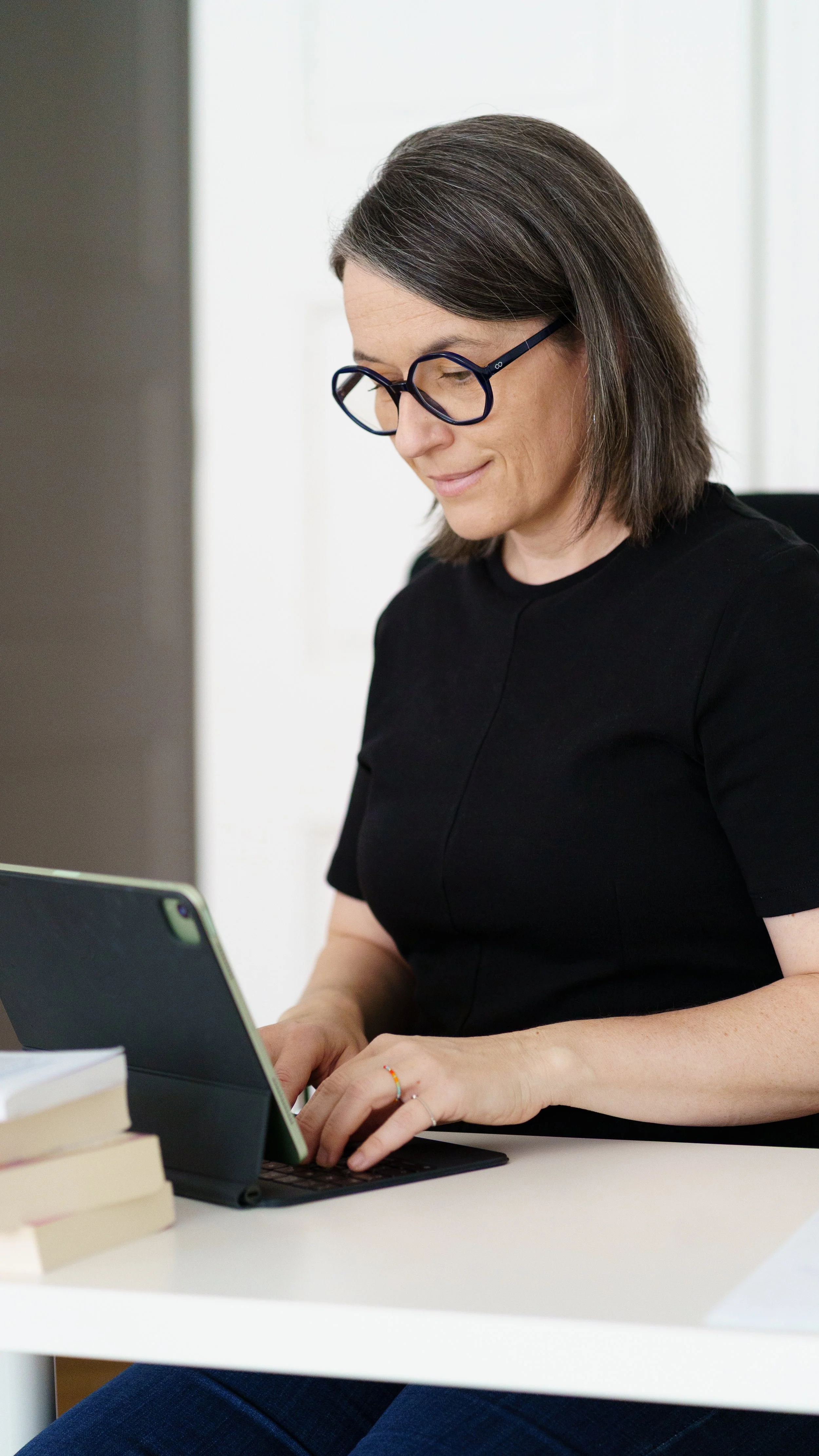 Eine Frau mit Brille sitzt an einem Schreibtisch und tippt auf einem Tablet. Es liegen Bücher auf dem Tisch.