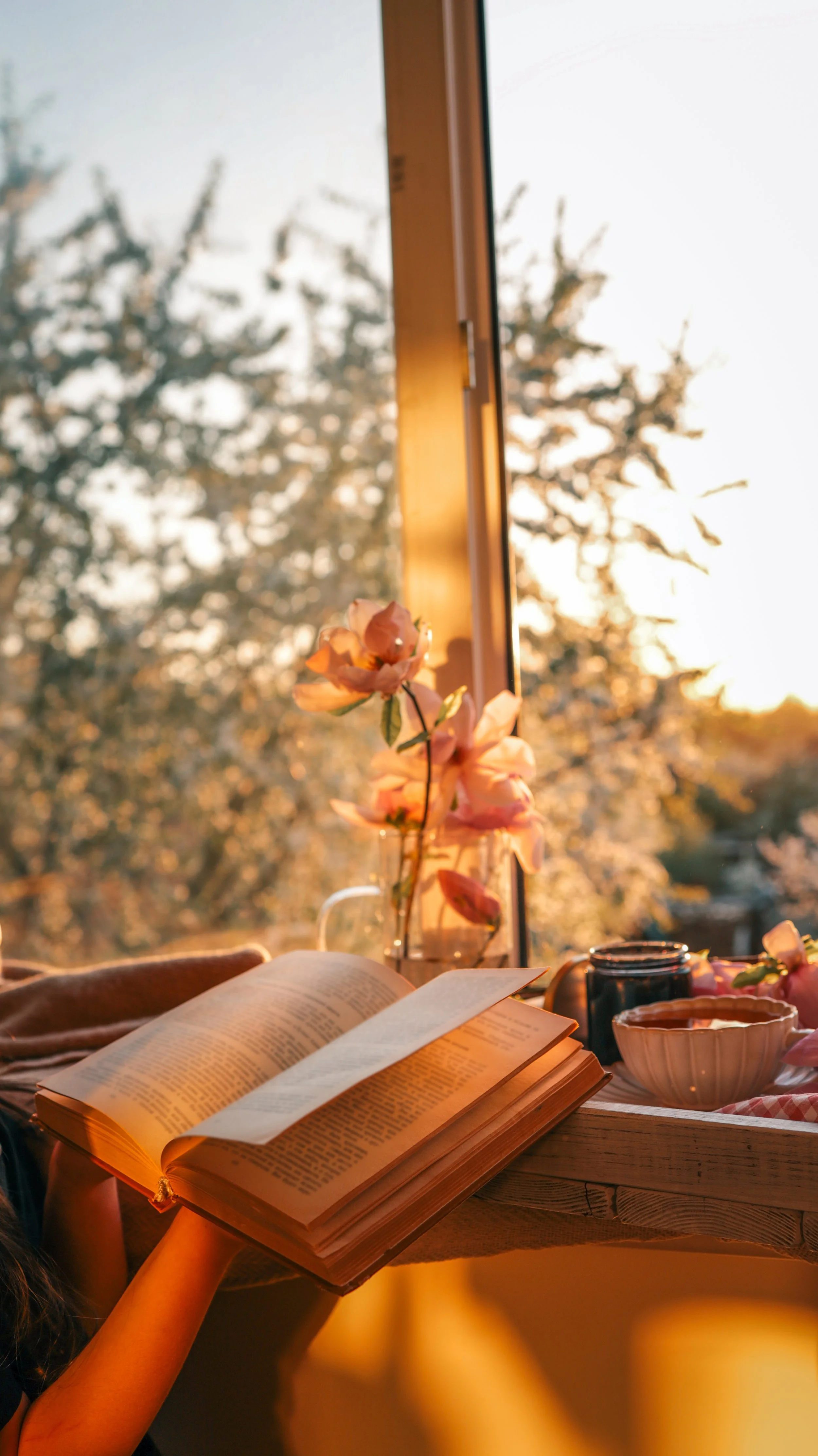 an open book and flowers and tea in front of an open window at dusk
