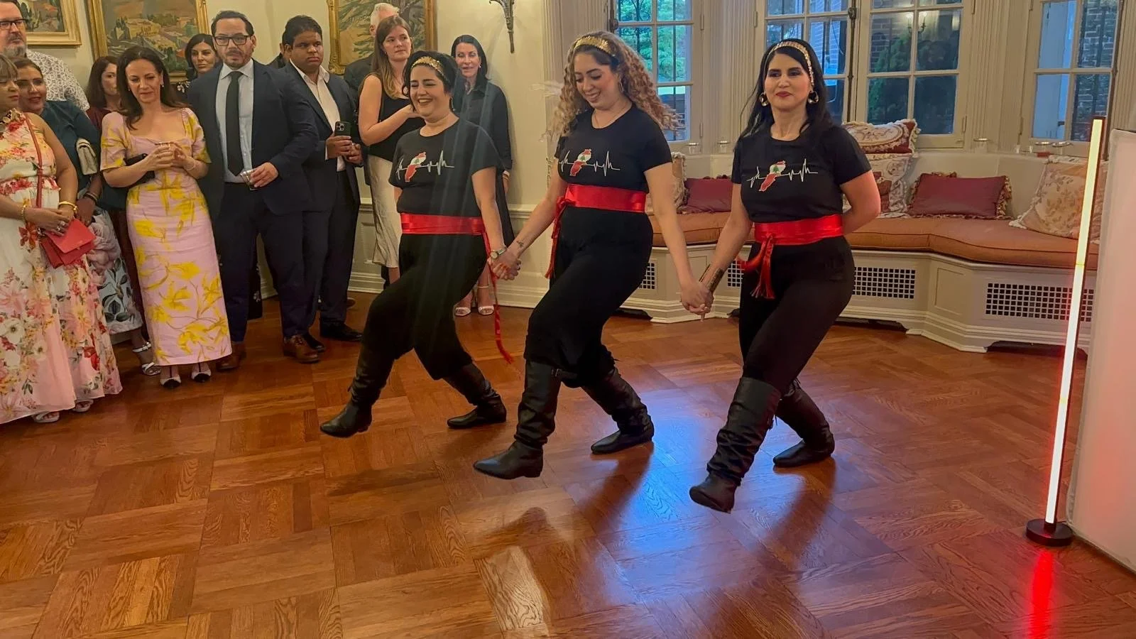 Three women dressed in black outfits with red sashes perform a traditional dance while holding hands in a room full of onlookers, some taking photos. The room has wooden floors, large windows, and ornate furniture.