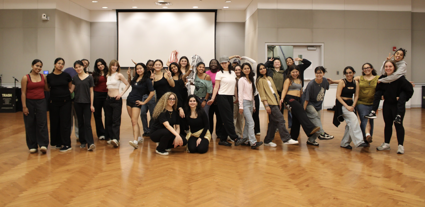 Group of young women standing and posing in a dance studio with wooden floors and a large projection screen in the background.
