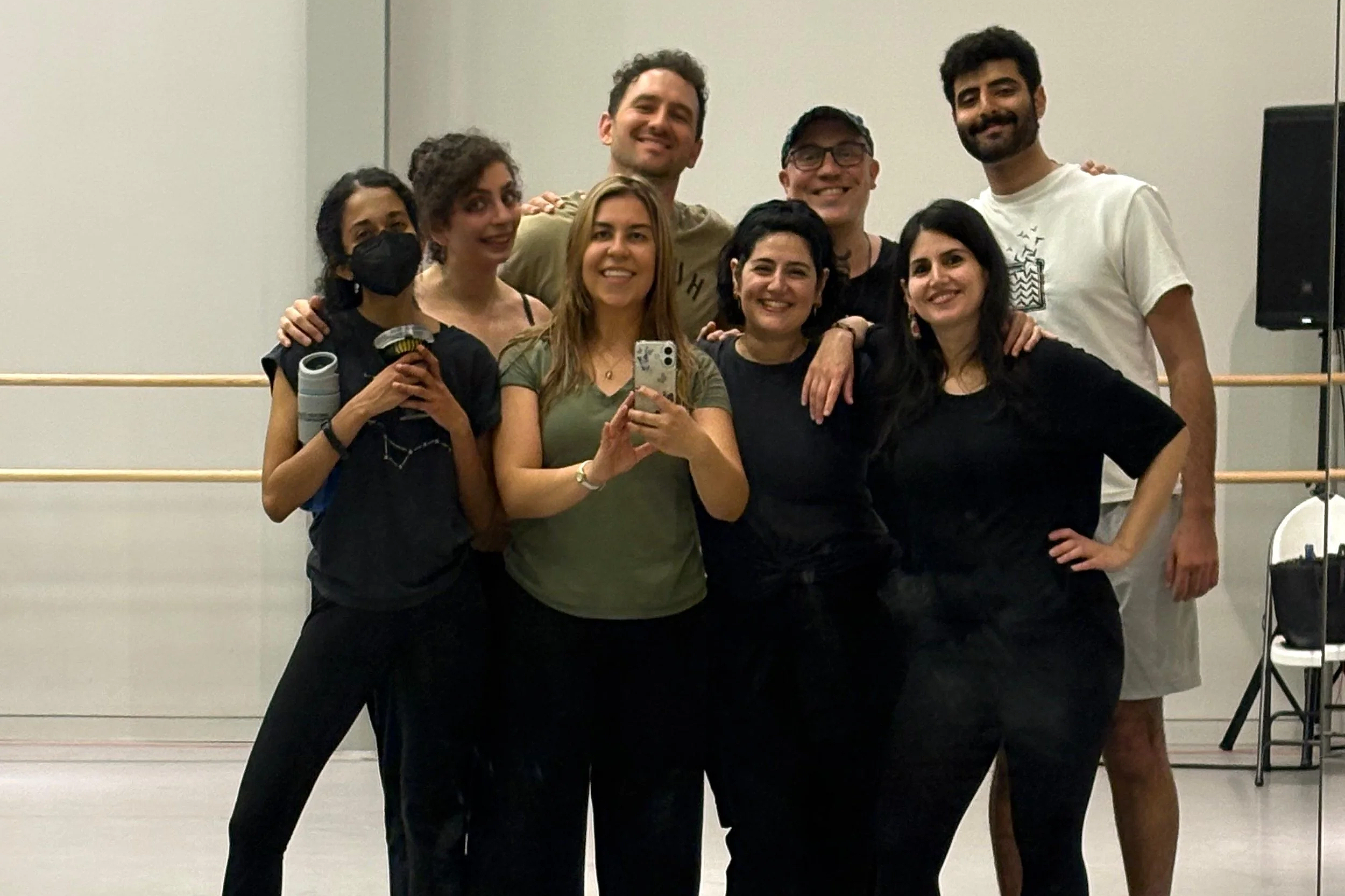 A group of nine diverse people posing together and smiling, with some making gestures, in a room with a plain background and a speaker on the right.