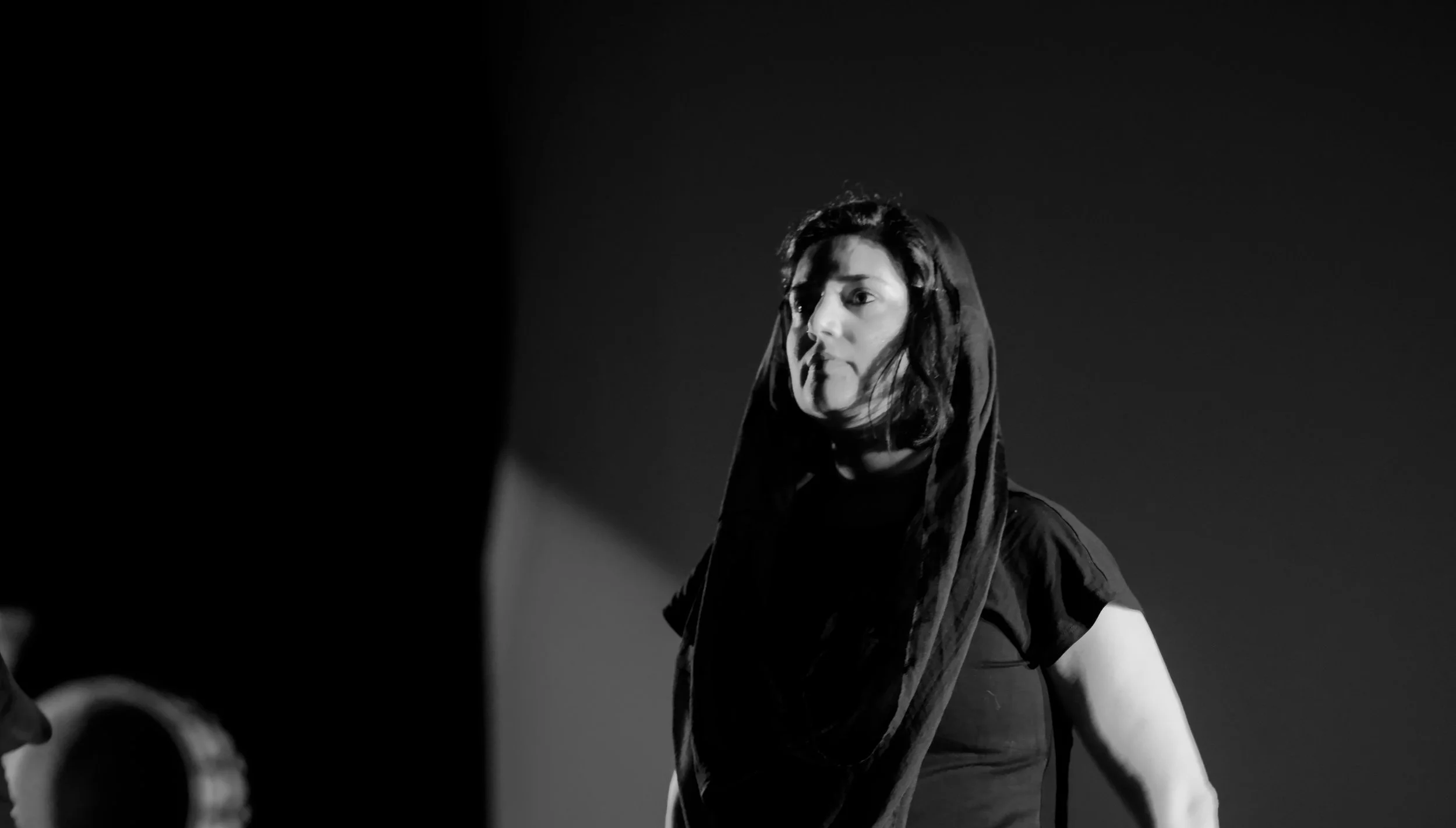 Black and white photo of a woman with long hair, wearing a hooded jacket, looking to the side against a dark background.