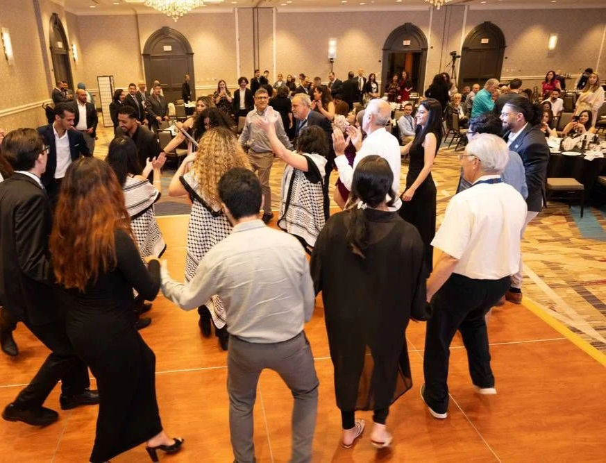 A group of people dancing in a circle at an indoor event with tables and attendees in the background.