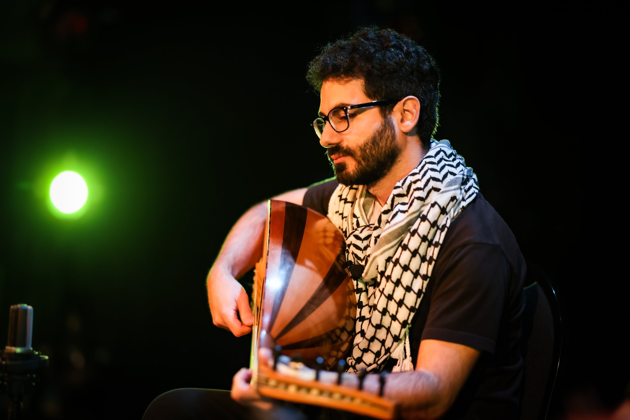 A man with dark curly hair, beard, glasses, and a patterned scarf is playing an acoustic guitar against a black background with a green light source to his left.