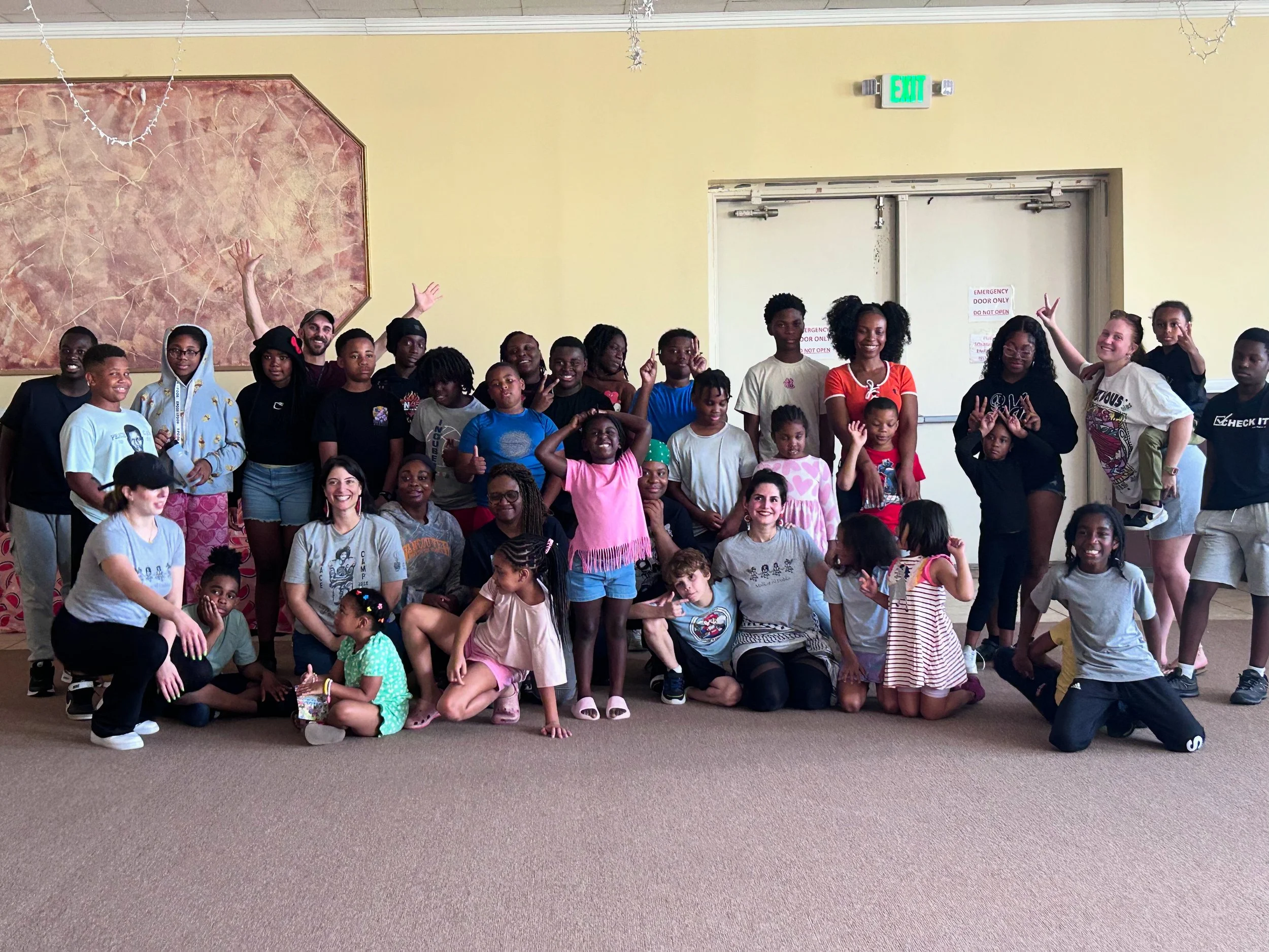 Group of children and adults posing together in a room, smiling and making peace signs, with a large painting on the wall and a door labeled 'Emergency Door Only' in the background.