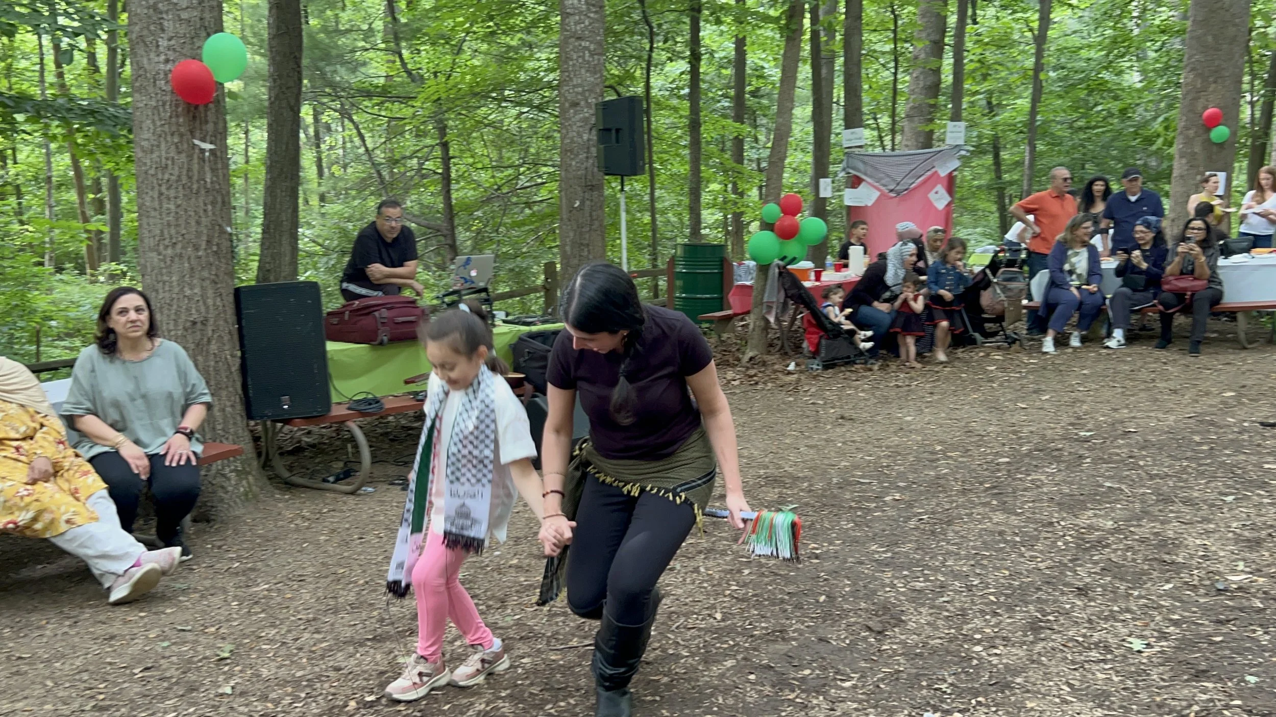 People attending an outdoor event in a forested area, decorated with red and green balloons. A woman and child are dancing in the foreground, while others sit and stand nearby, enjoying the gathering.